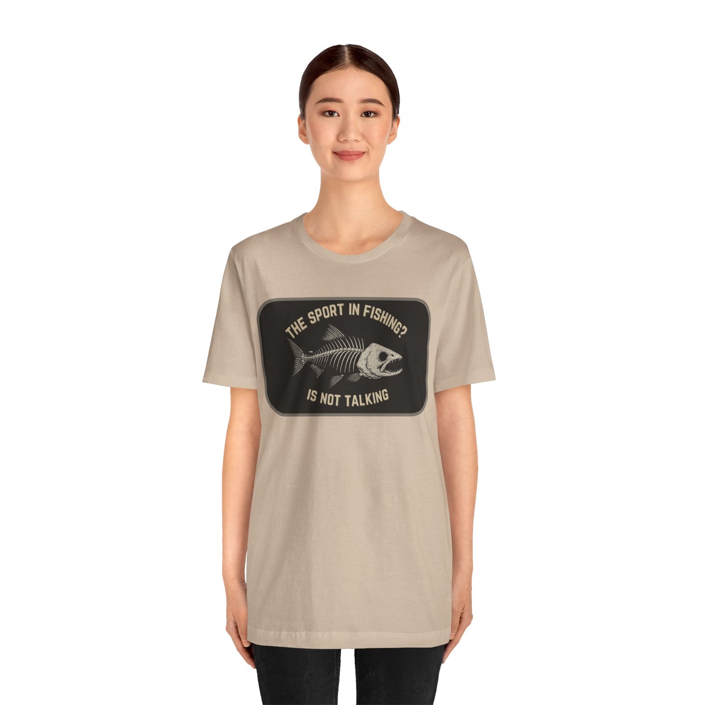 A woman faces forward in a plain white setting, wearing Printifys The Sport in Fishing? Tee—a beige unisex shirt featuring a distressed fish skeleton print and the text: THE SPORT IN FISHING? IS NOT TALKING.