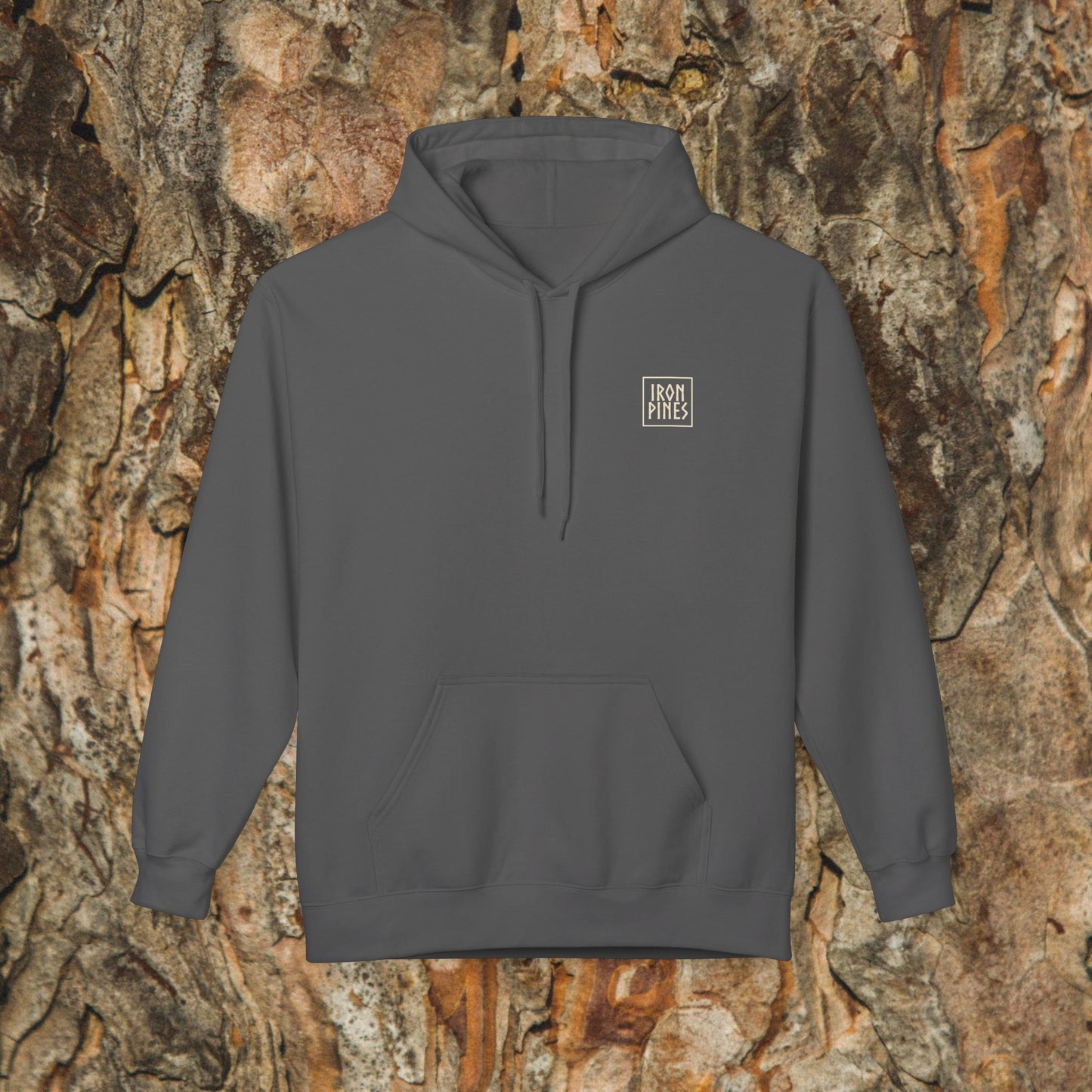 The Iron Pines Hoodie by Printify, in charcoal gray with a small white chest logo, is sustainable outdoor apparel shown against textured tree bark.