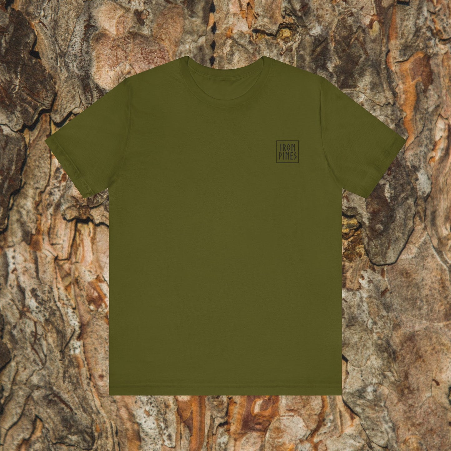 The Iron Pines Crest Tee by Printify features an olive green Airlume cotton shirt with a small black square IRON PINES logo on the left chest, shown against textured tree bark for a stylish and sustainable look.
