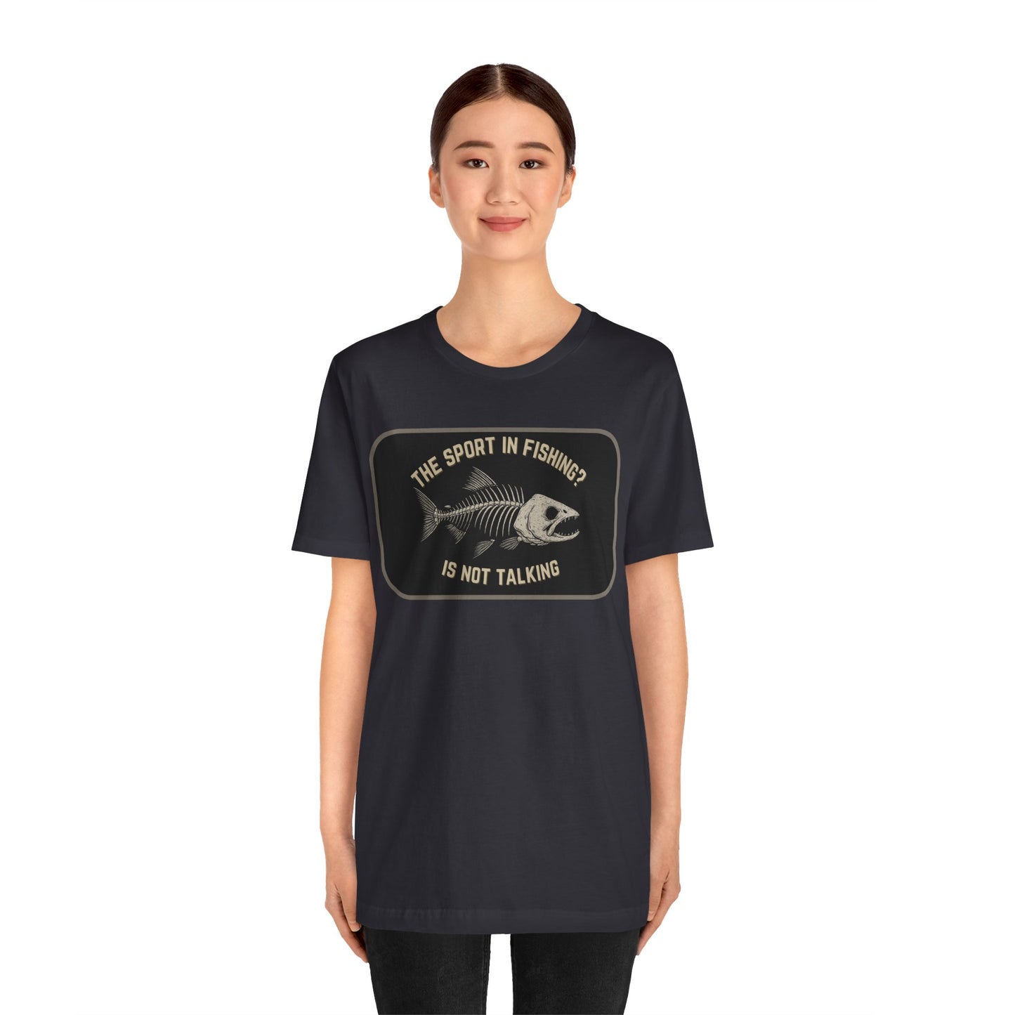 A person faces forward in a black Printify The Sport in Fishing?” Tee, featuring a distressed fish skeleton graphic and the text The sport in fishing? Is not talking. The background is plain white.
