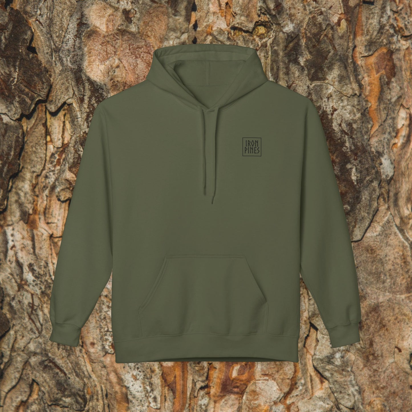 The Iron Pines Hoodie by Printify, in olive green midweight fleece with a small left-chest IRON PINES logo, is shown against textured tree bark—a sustainable choice for outdoor adventures.