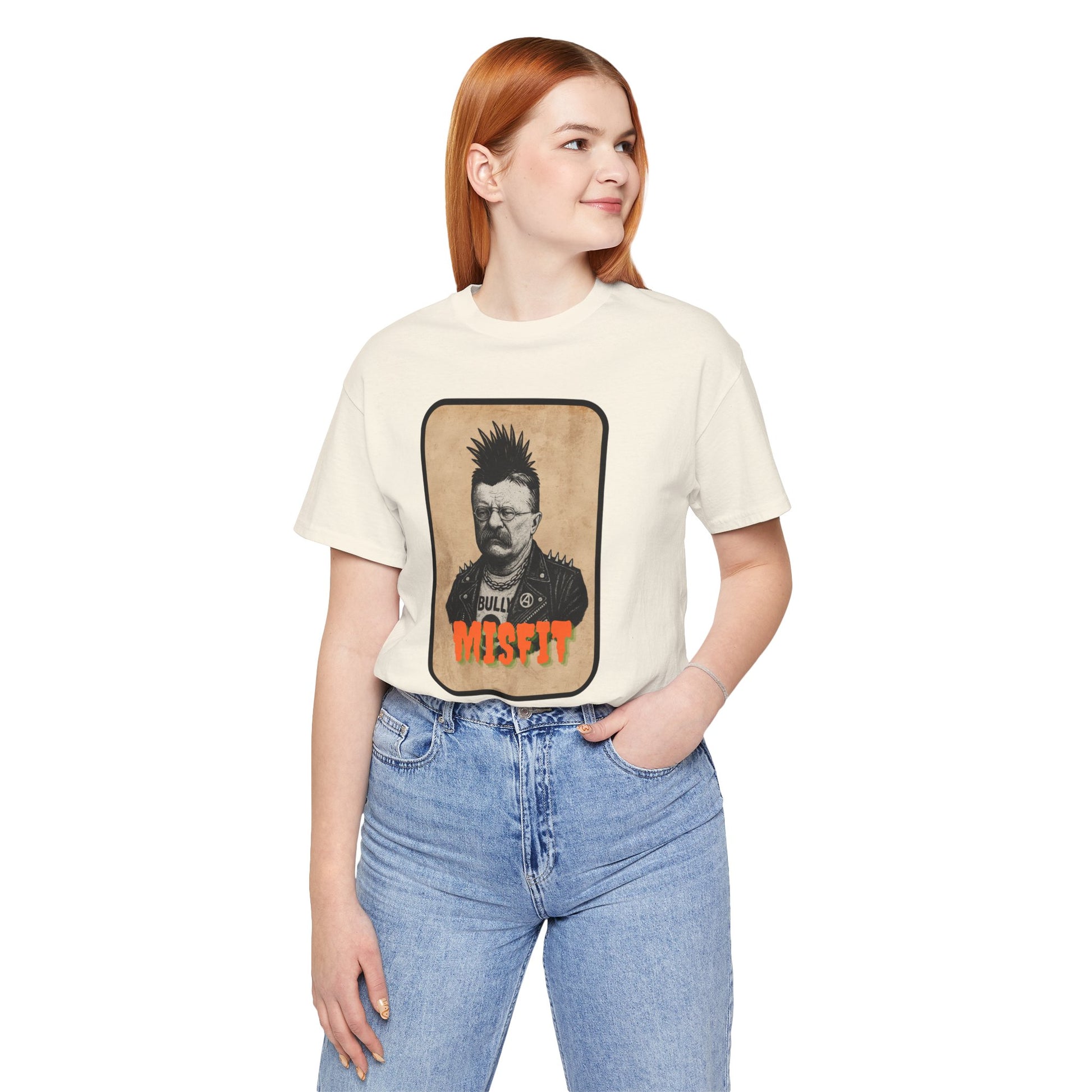 A woman with long red hair, wearing the Iron Pines Teddy Roosevelt Punk T-Shirt from Misfit History, stands smiling with one hand in her pocket. She pairs the white rebel-inspired tee with light blue jeans for a relaxed look.