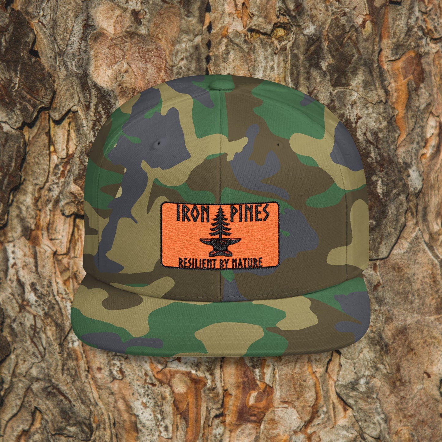 The Iron Pines Logo Hat by Printify is a camouflage snapback with an embroidered patch displaying a pine tree and the text IRON PINES RESILIENT BY NATURE on a textured tree bark background.