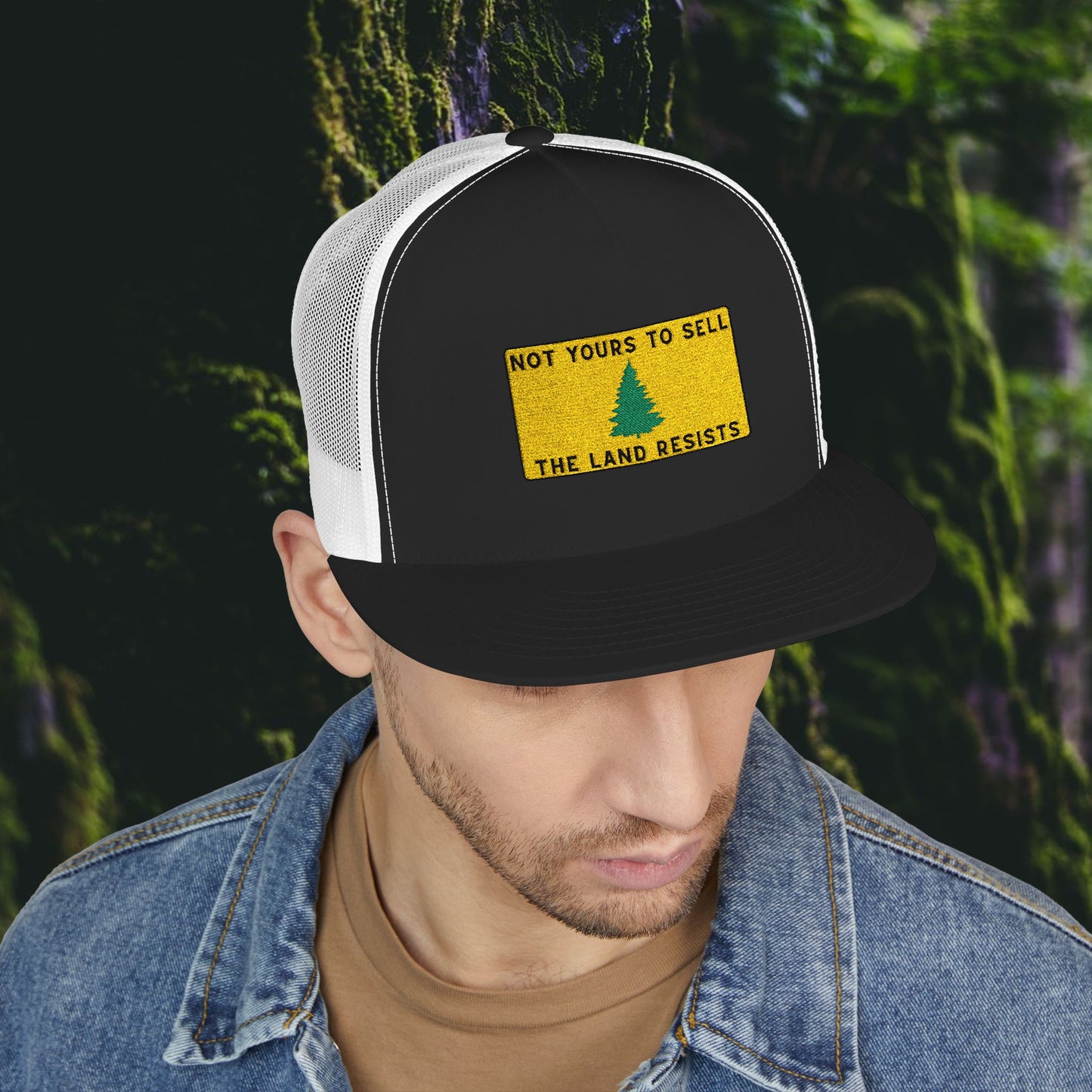Wearing the Printify Public Land Trucker Hat—black and white, reading NOT YOURS TO SELL THE LAND RESISTS above a green tree—a person stands against a blurred backdrop of greenery and moss.