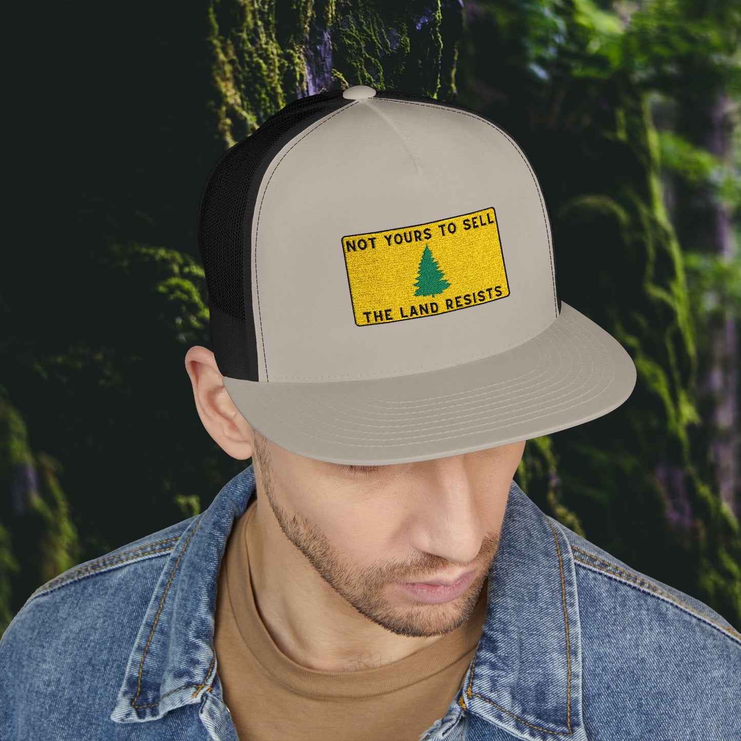 A man in a denim jacket and brown shirt looks down, wearing the Printify Public Land Trucker Hat—beige and black with a yellow NOT YOURS TO SELL THE LAND RESISTS patch. This hat highlights environmental activism.