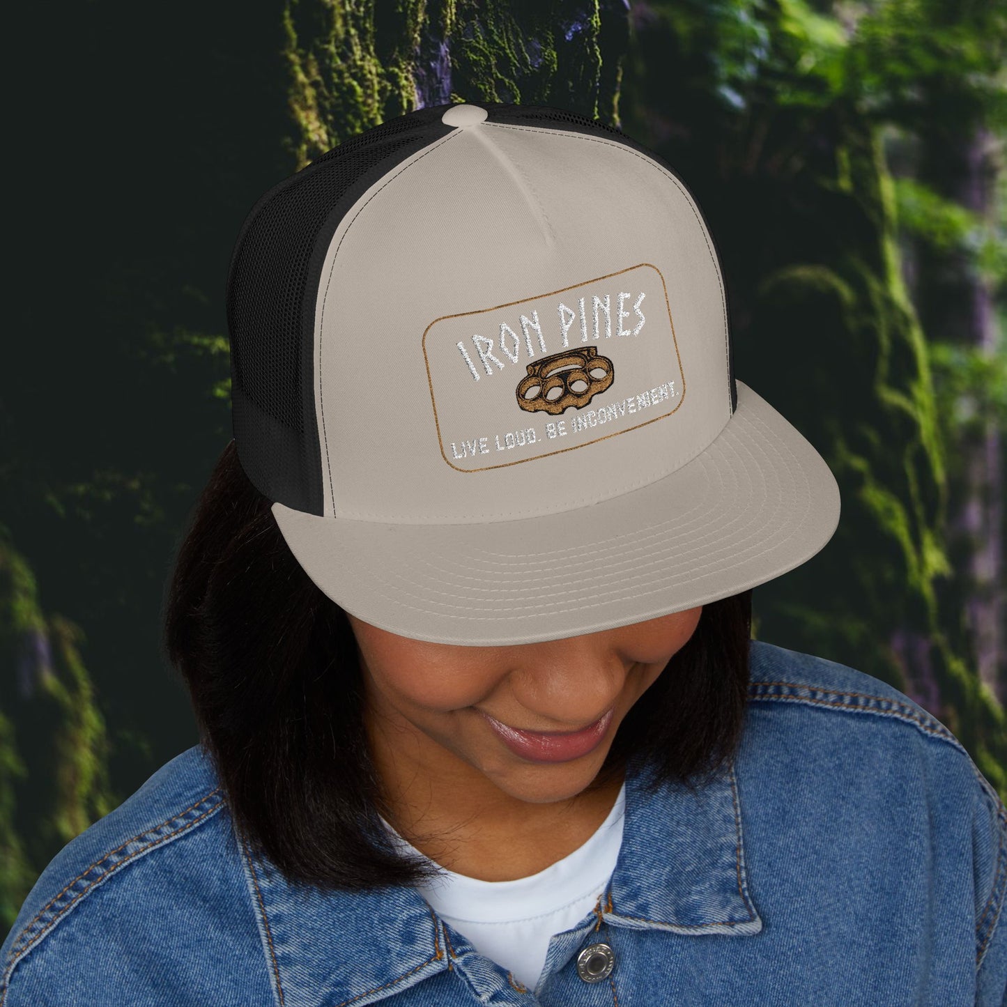 A person in a denim jacket and the Printify Iron Pines Trucker Hat – “Live Loud. Be Inconvenient.” Brass Knuckle Edition stands before a blurred forest, smiling while looking down.