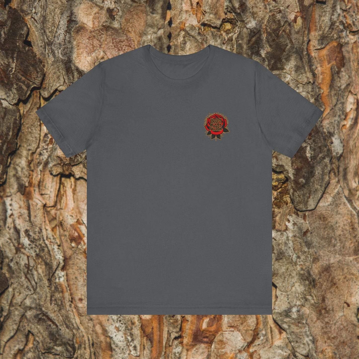The Never Mistake My Kindness For Weakness Tee by Printify is a unisex charcoal gray graphic t-shirt with a small red rose and text on the left chest, shown against a tree bark background.