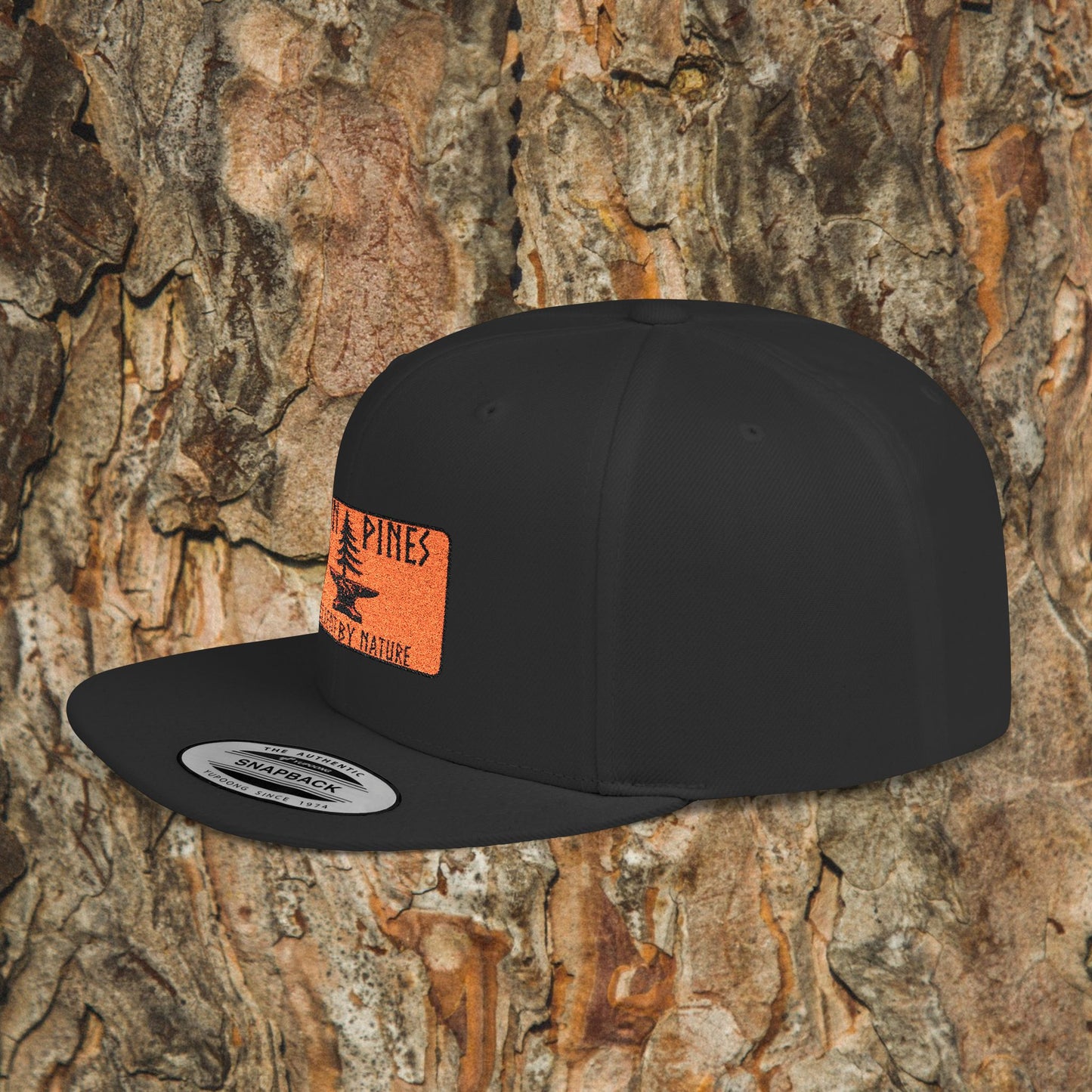 The Iron Pines Logo Hat by Printify, a black structured snapback with an orange rectangular patch featuring the Iron Pines Co logo and tree graphic, is shown against textured tree bark.