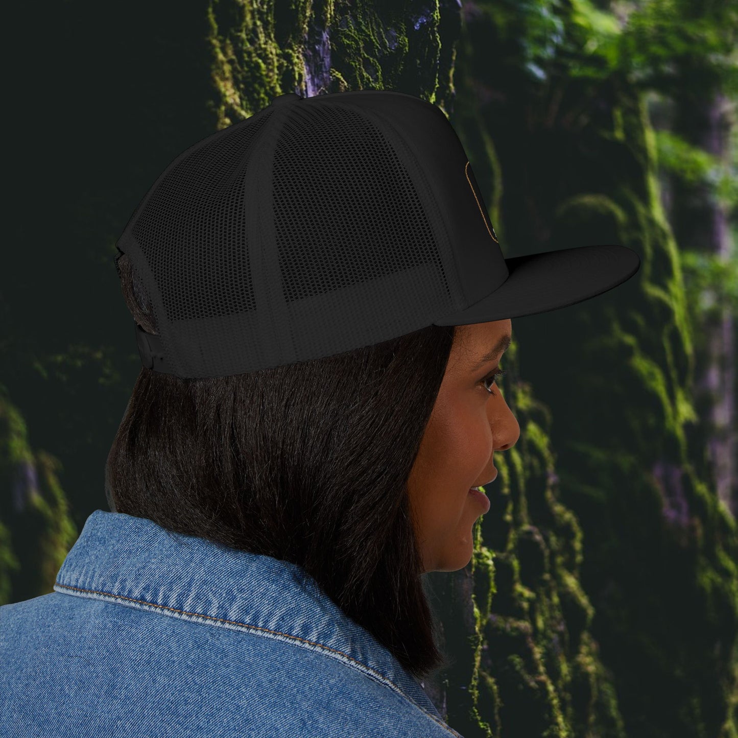 A woman with straight dark hair wears the Printify Iron Pines Trucker Hat – “Live Loud. Be Inconvenient.” Brass Knuckle Edition and a denim jacket, shown in profile against a blurred green, mossy outdoor background.