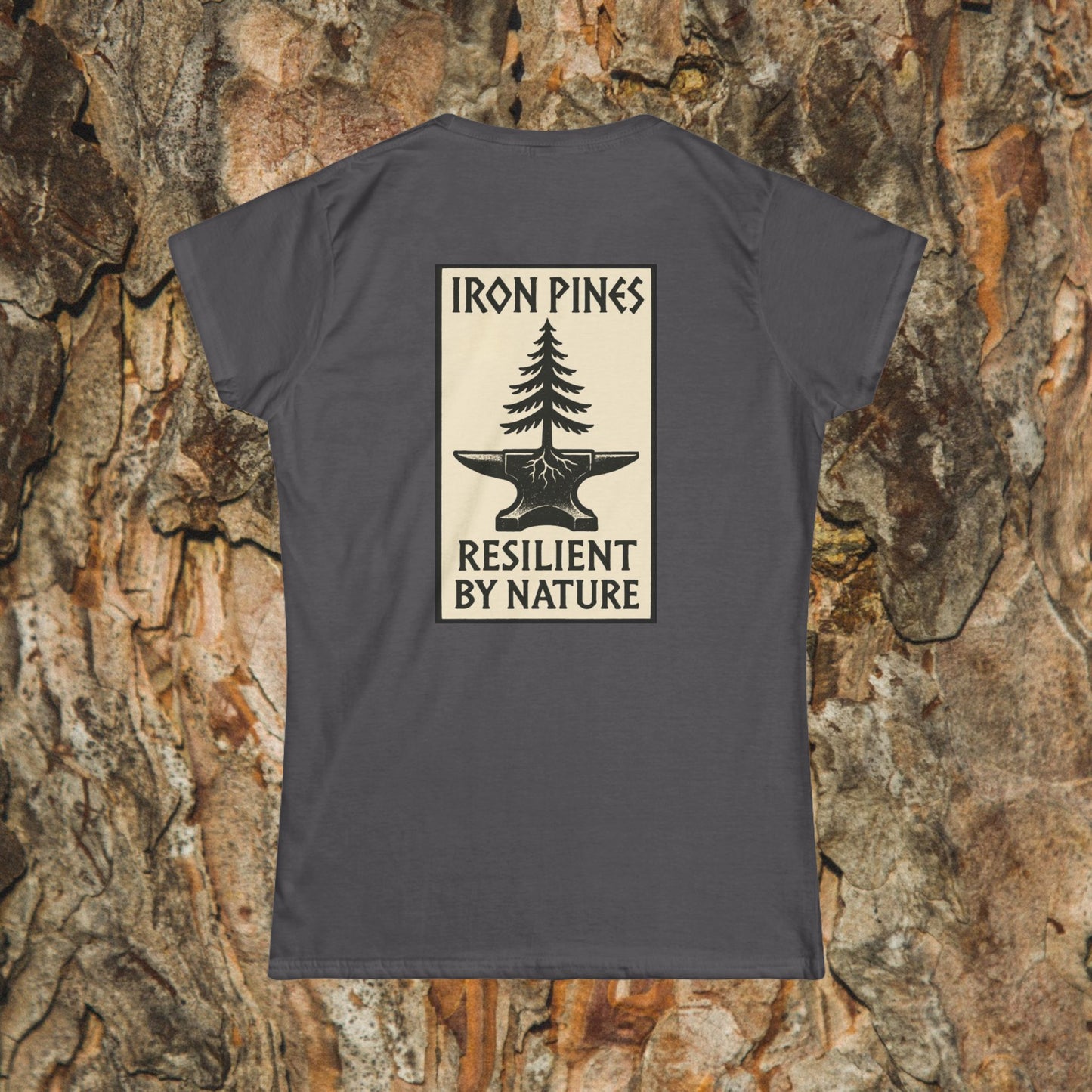 The Iron Pines Womans Tee by Printify features a women’s softstyle fit with a rectangular pine tree on an anvil graphic, “IRON PINES” above and “RESILIENT BY NATURE” below, set against a bark background.