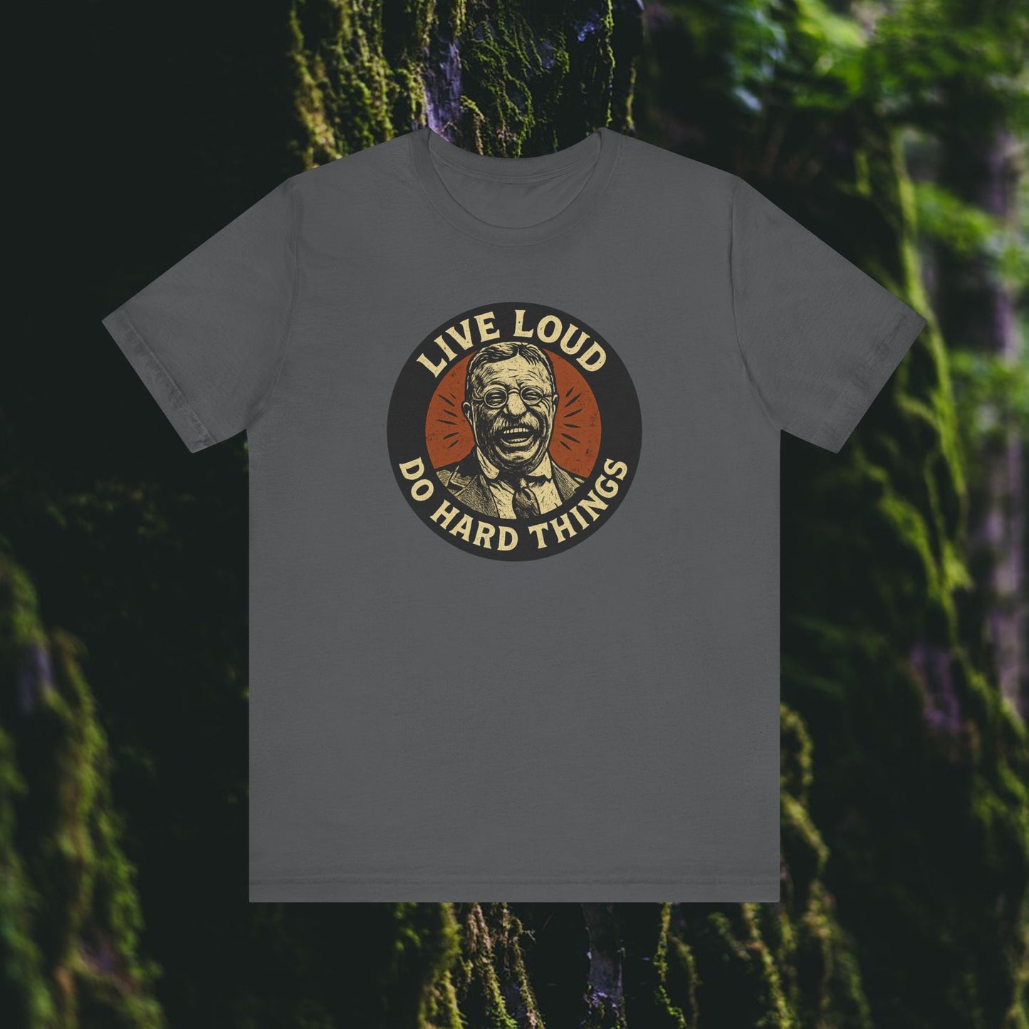 The Live Loud, Do Hard Things Teddy Roosevelt Tee by Printify features a circular graphic of Teddy with glasses, the bold mottos, and a mossy green tree background—perfect for those who seek inspiration and adventure.