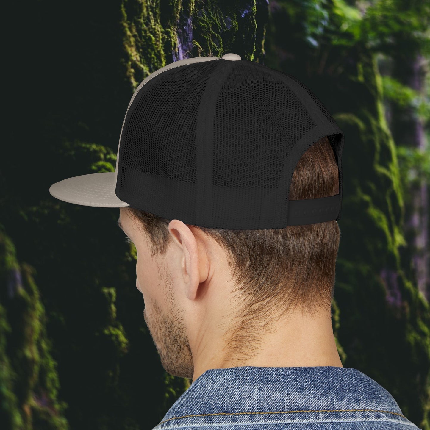 A person wearing the Printify Public Land Trucker Hat, Not Yours To Sell The Land Resists, and a blue denim jacket stands with their back to the camera against a lush, mossy backdrop.