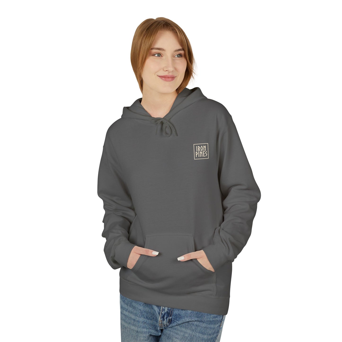 A woman with light brown hair wears The Iron Pines Hoodie by Printify, a sustainable gray hoodie with a small chest logo. She faces sideways, smiling slightly, hands in the front pocket, paired with blue jeans.