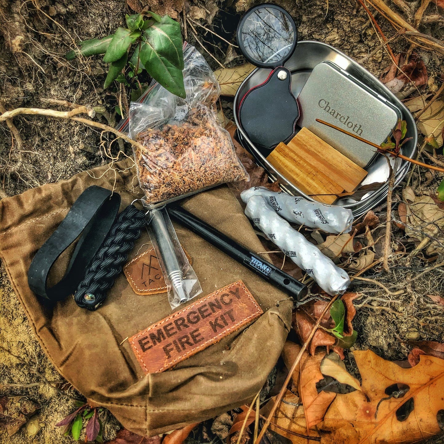 A TITAN Survival TACAMO 15-Piece Emergency Fire Kit with a brown waxed canvas bag sits on the ground, featuring fire starter tools like a ferro rod, paracord, fatwood, charcloth tin, magnifier, and tinder among autumn leaves.
