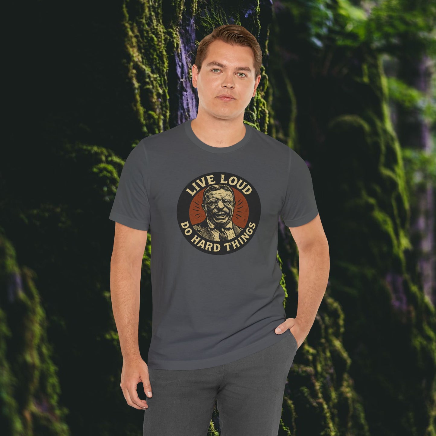 A man stands before a mossy wall, wearing Printify’s Live Loud, Do Hard Things Teddy Roosevelt Tee, featuring a bold face graphic and motto. He looks confidently at the camera with one hand in his pocket.