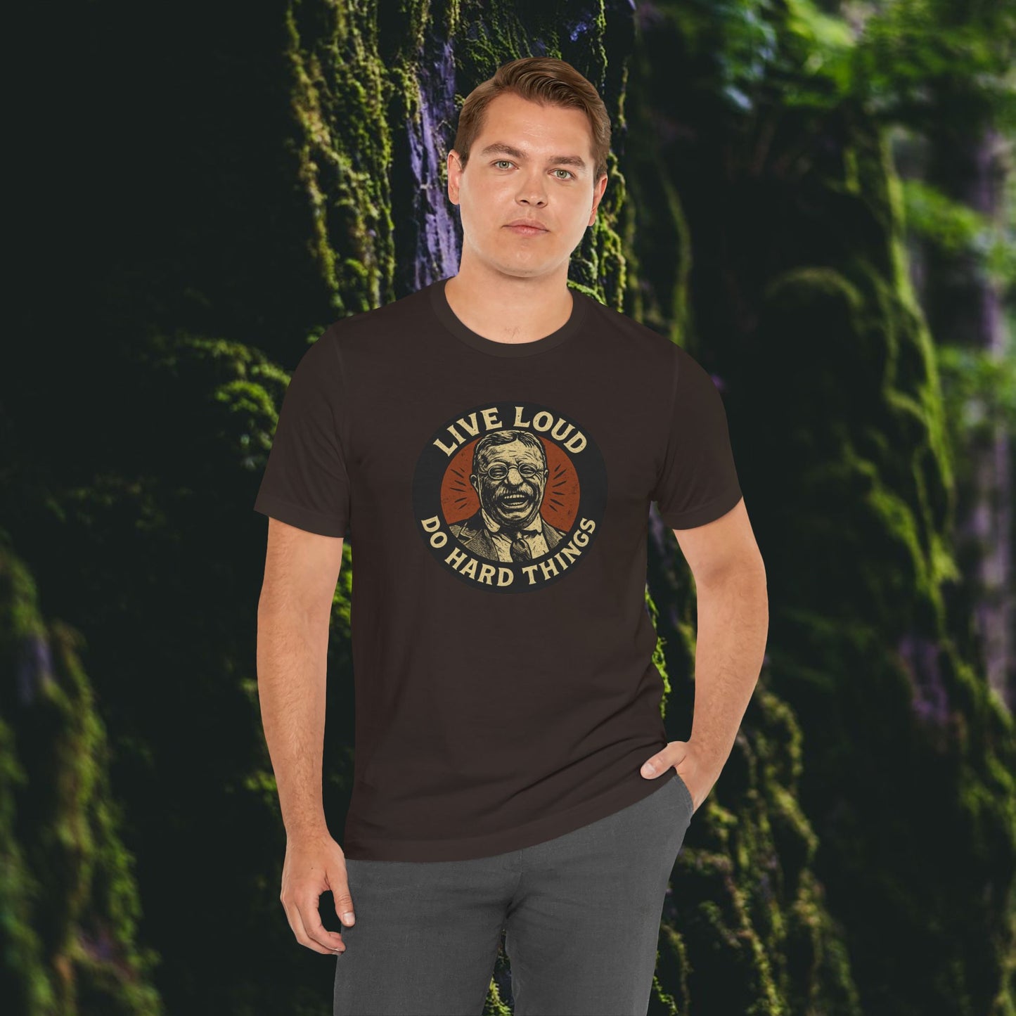 A man stands in a mossy forest wearing Printifys Live Loud, Do Hard Things Teddy Roosevelt Tee, which features an illustrated face of Roosevelt and bold retro-style lettering.