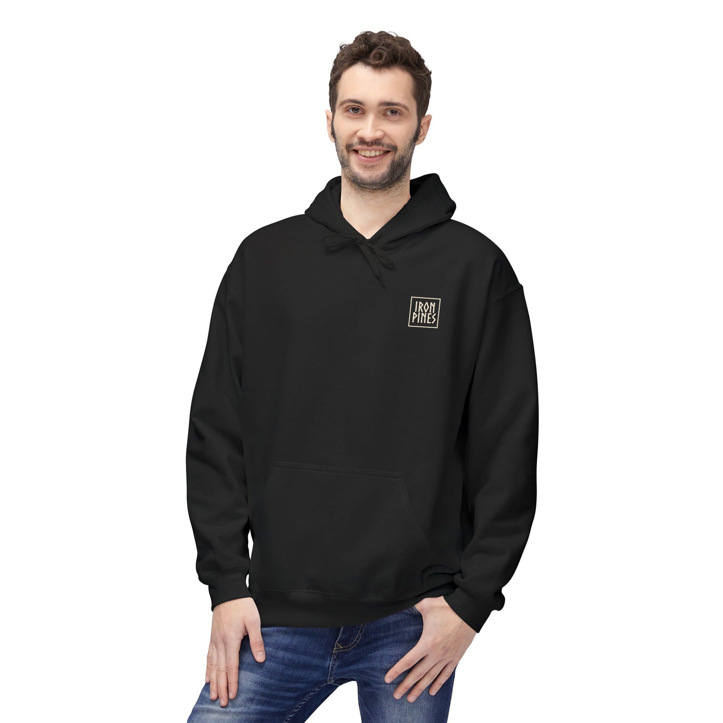 A smiling man with short brown hair and a beard wears The Iron Pines Hoodie by Printify, made from midweight fleece with a small white left chest logo, paired with blue jeans, standing against a white background.