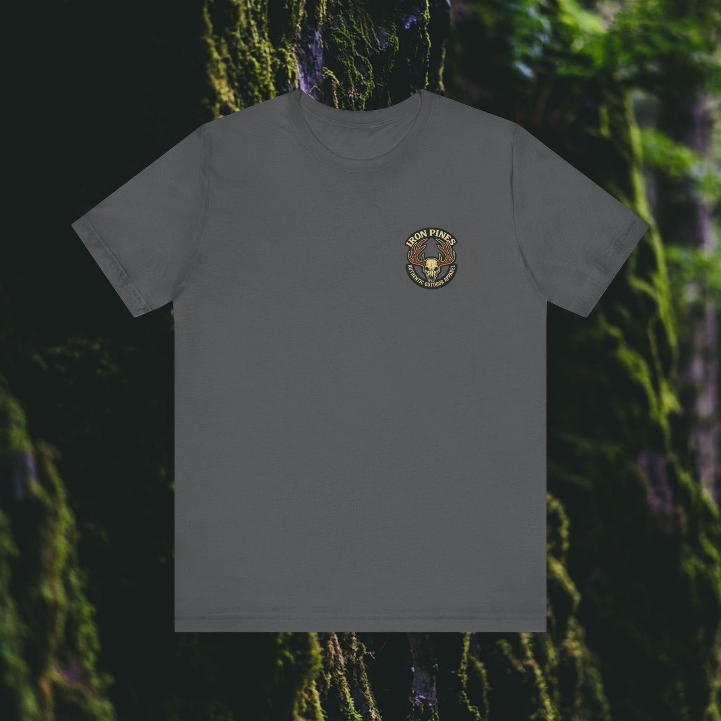 The I Hunt Because Bail’s Expensive Tee by Printify is a durable gray outdoor shirt featuring a small Iron Pines logo—pine tree and axe—on the left chest, set against mossy forest trees in the background.