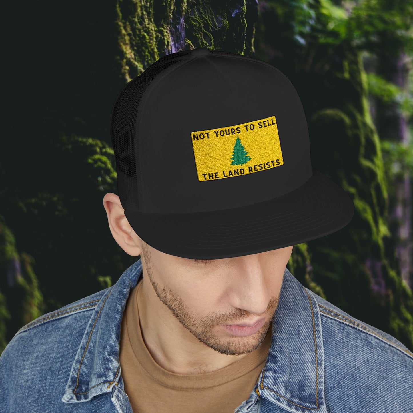 Wearing the Printify Public Land Trucker Hat—a black snapback with a yellow NOT YOURS TO SELL, THE LAND RESISTS patch and green tree—a person in a denim jacket over a brown shirt stands before mossy trees.