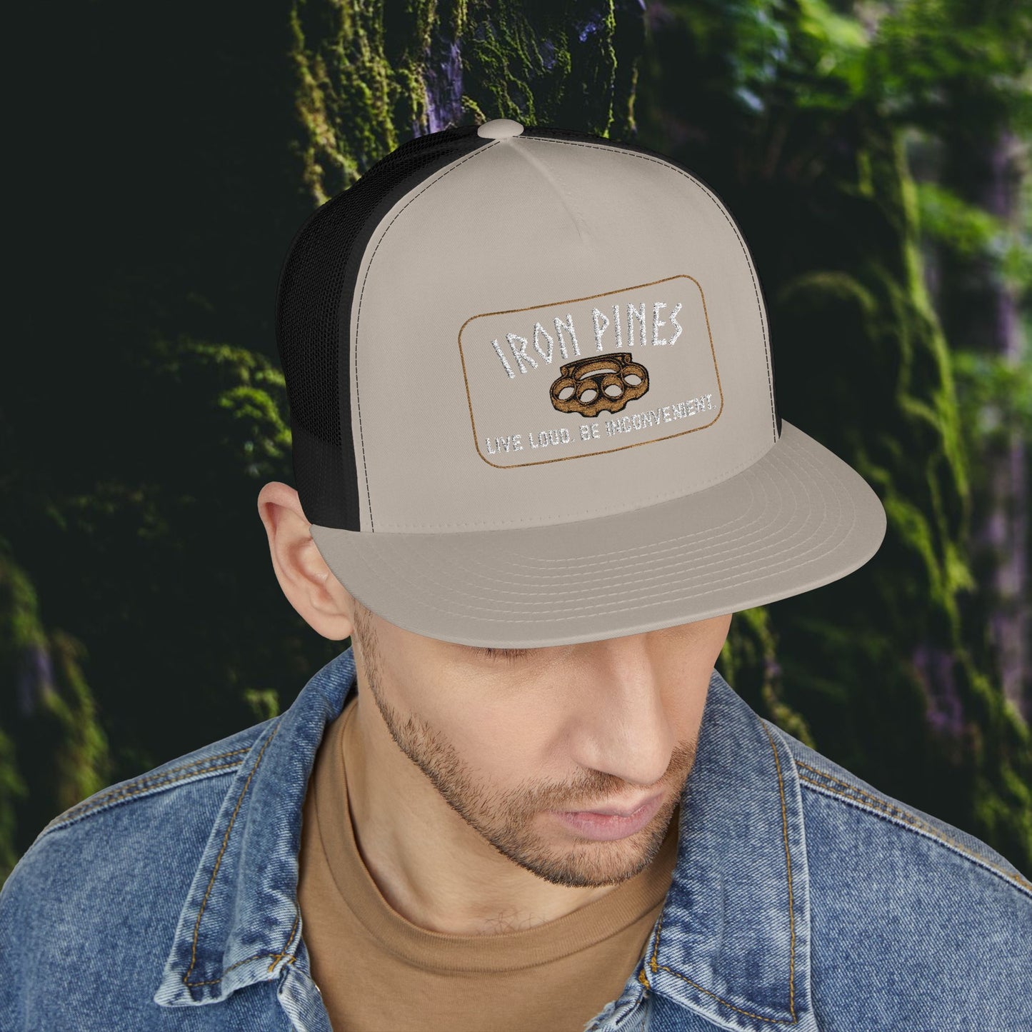 A man wears the Printify Iron Pines Trucker Hat – “Live Loud. Be Inconvenient.” Brass Knuckle Edition, in beige and black, with brass knuckles embroidery. He looks down by tree bark with green moss in the background.
