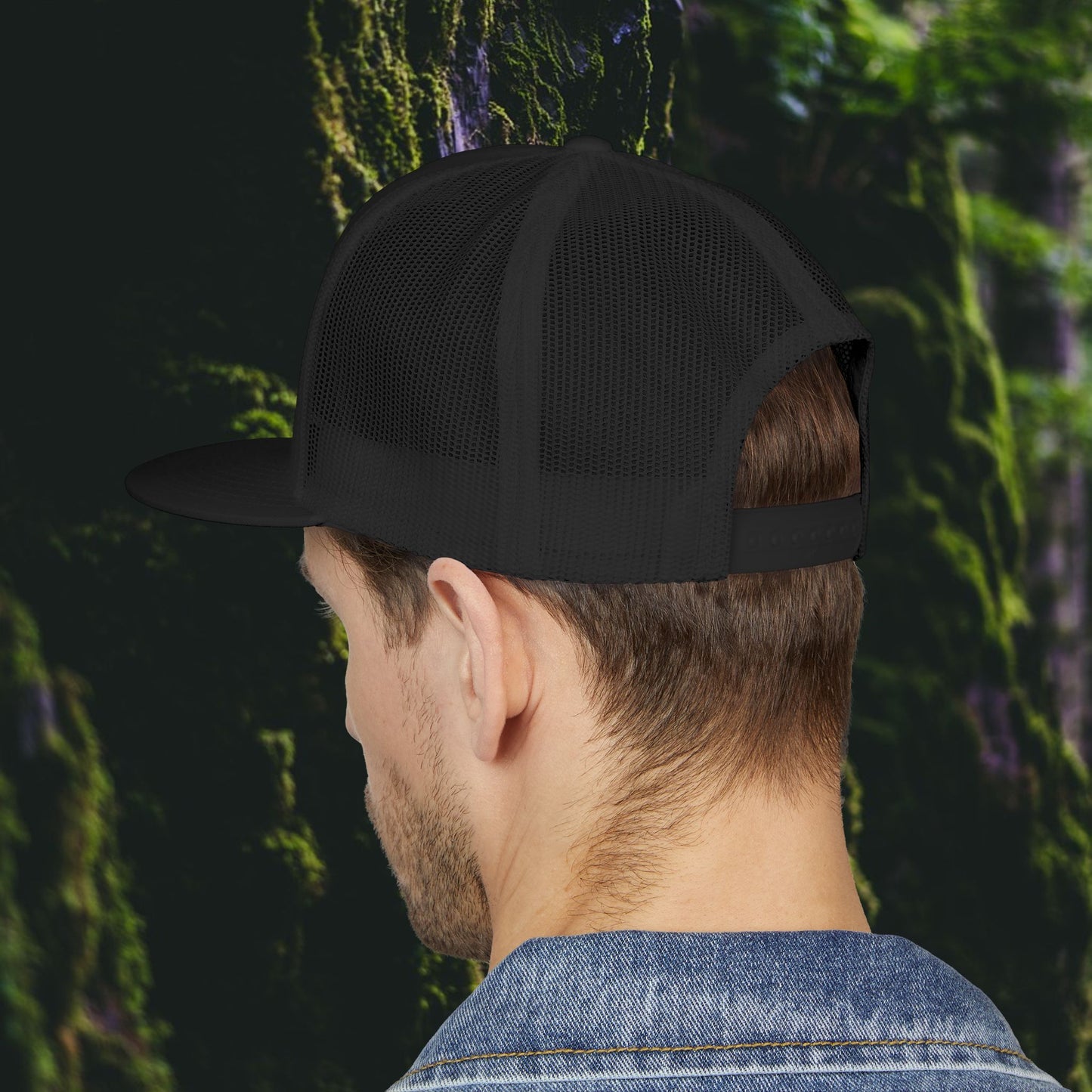 A man in a Printify Public Land Trucker Hat, Not Yours To Sell The Land Resists, and a denim jacket faces away before moss-covered rocks.