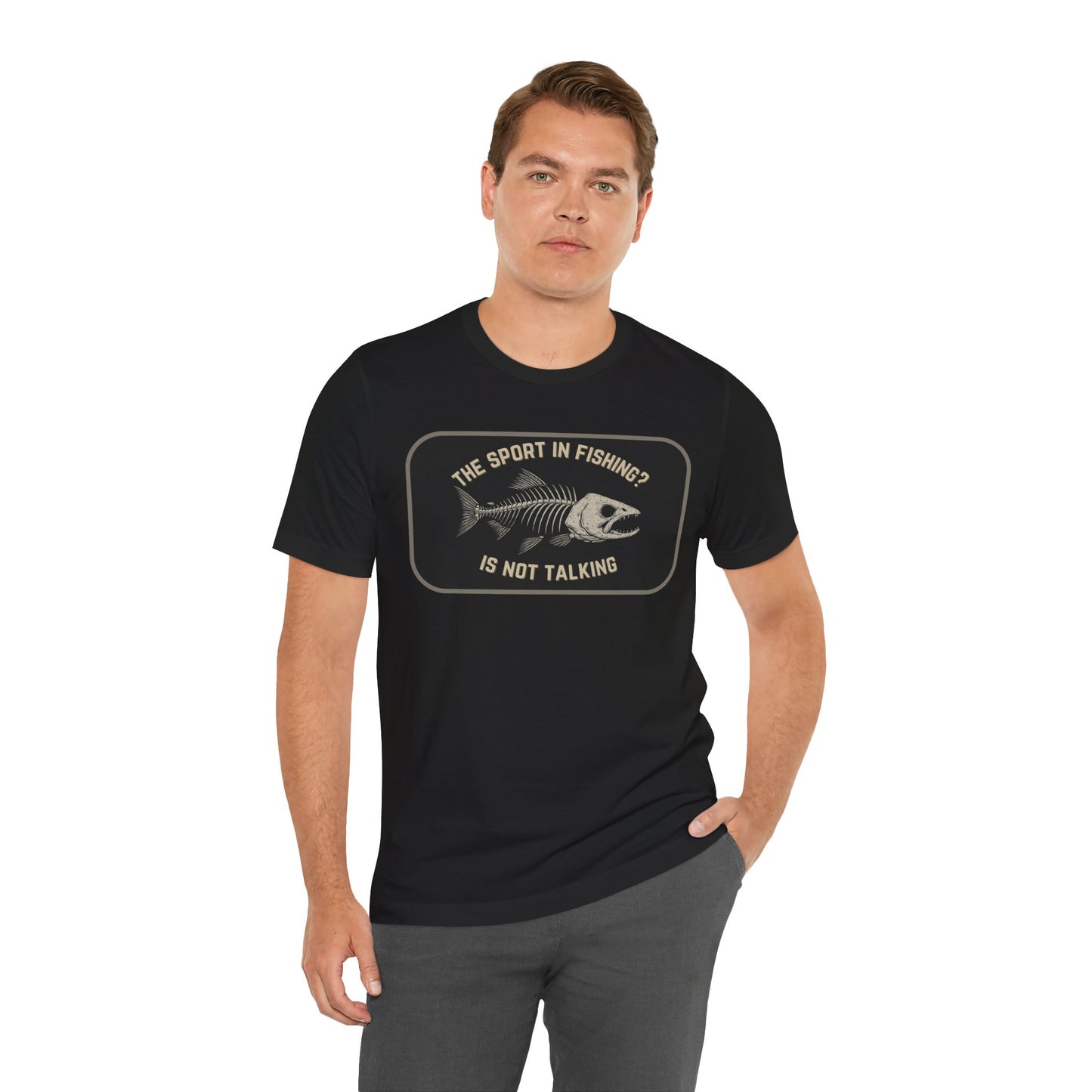 A man wears Printify’s “The Sport in Fishing?” Tee, a black unisex shirt with a distressed fish skeleton and “The sport in fishing? Is not talking” text, posing with one hand in his pocket and looking at the camera.