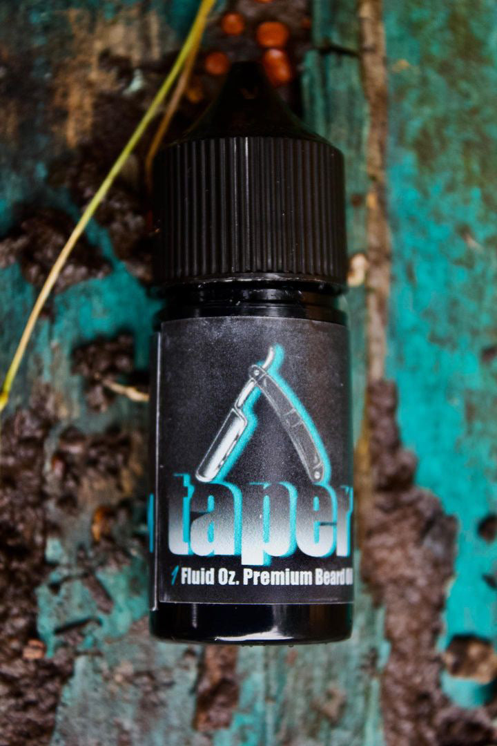 A small bottle of TAPER Beard Oil & Balm by Bedlam Beard Company in cedar leather scent, with a straight razor image and Fluid Oz. text, rests on a rustic wooden surface with turquoise and brown textures.