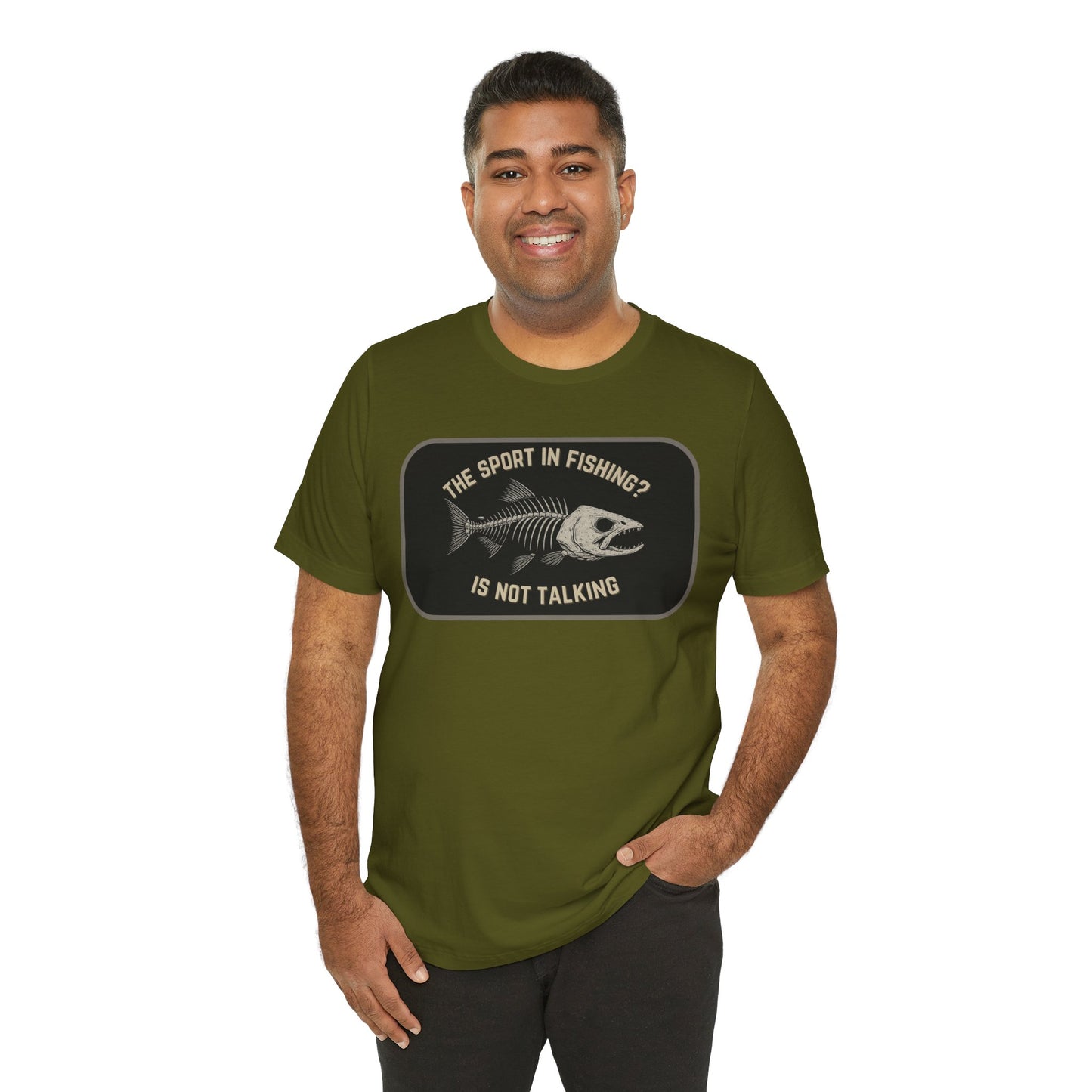 A smiling man with one hand in his pocket wears Printify’s The Sport in Fishing?” Tee—an olive green unisex shirt with a distressed fish skeleton print and the phrase, The sport in fishing? is not talking.