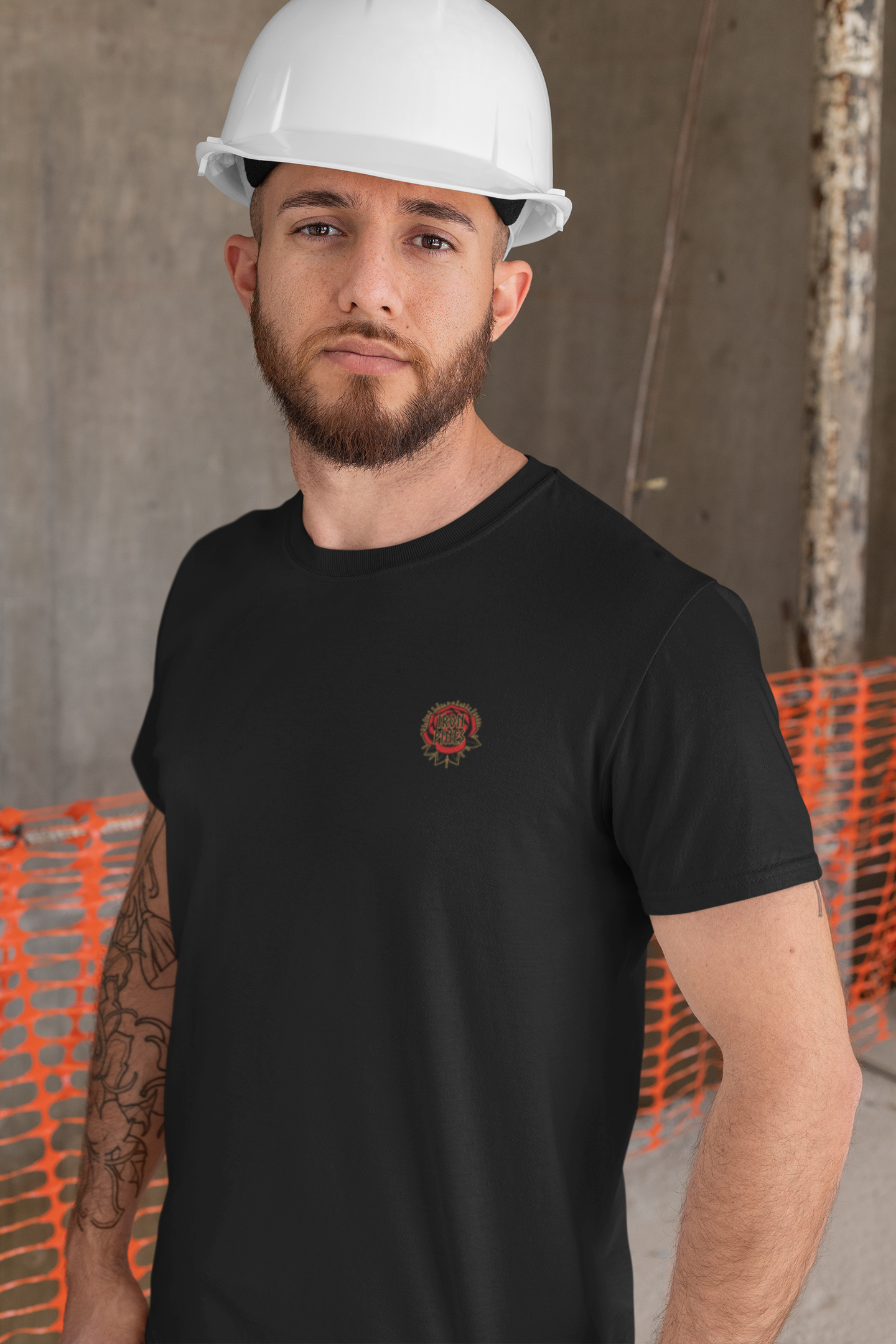 A bearded man in a Printify Never Mistake My Kindness For Weakness Tee and a white hard hat stands at a construction site, with orange safety netting and concrete walls behind him.
