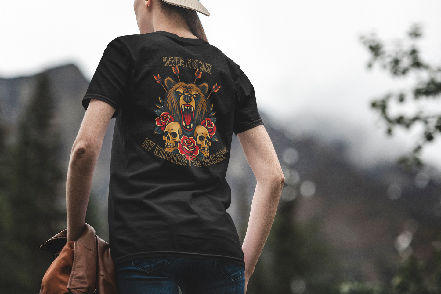 Outdoors, a person wears the Printify Never Mistake My Kindness For Weakness Tee—an all-gender graphic shirt displaying a roaring bear, roses, skulls, and bold text. Mountains and trees blur in the background.
