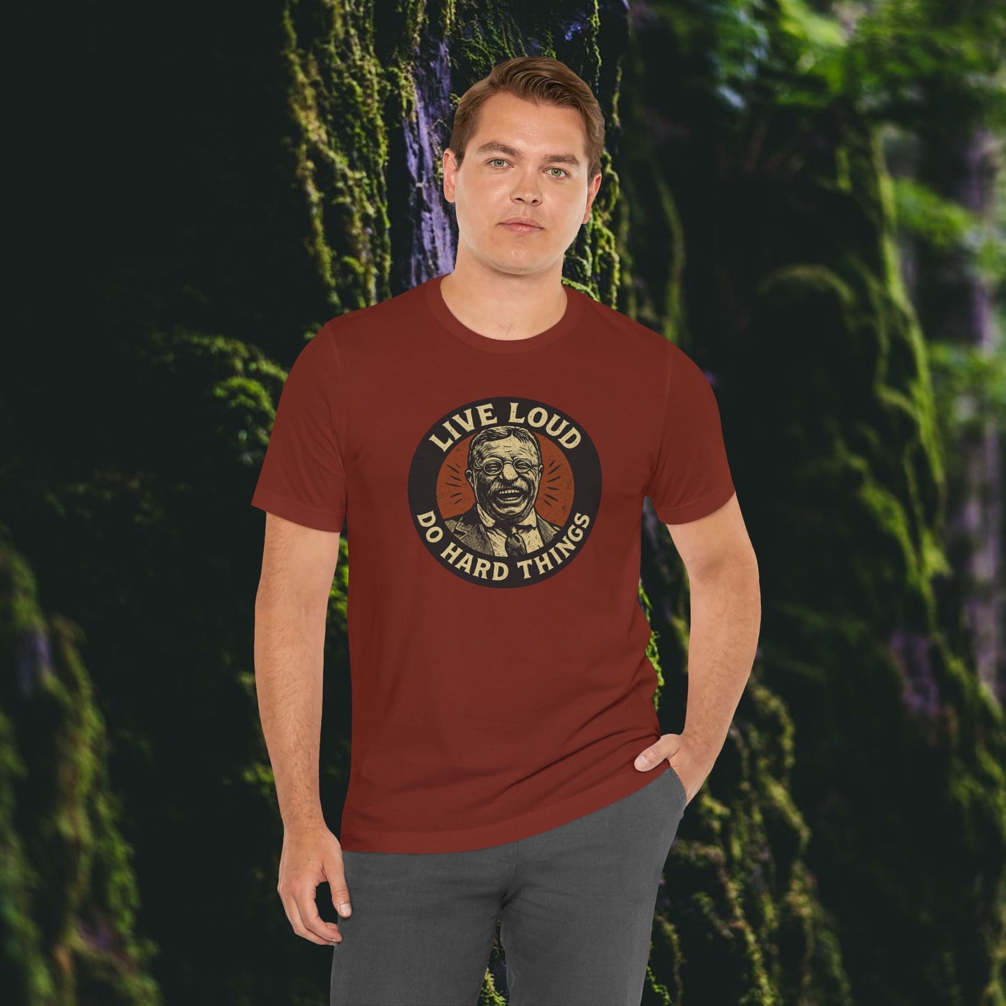 A man in a red Live Loud, Do Hard Things Teddy Roosevelt Tee by Printify, featuring an inspiring graphic of Teddy Roosevelt, stands before a lush, moss-covered wall with greenery in the background.