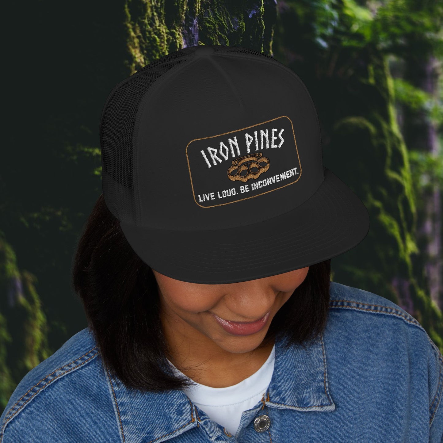 A woman smiles as she looks down, wearing the Iron Pines “Live Loud. Be Inconvenient.” Brass Knuckle Edition Trucker Hat by Printify with a blue denim jacket, surrounded by mossy rocks and greenery.