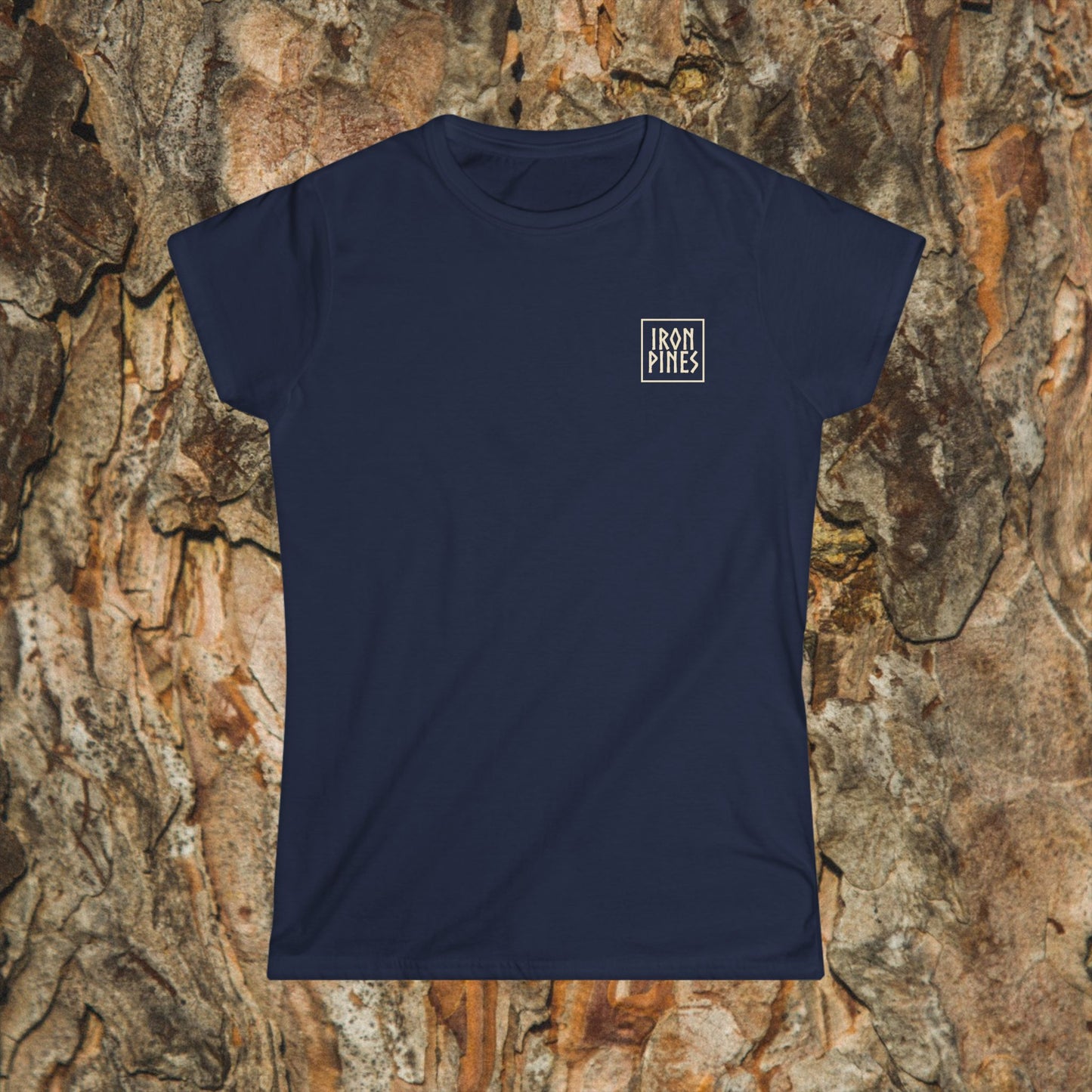 The Iron Pines Womans Tee by Printify in navy blue features a small white logo on the upper left chest. Displayed on tree bark, this softstyle tee blends comfort, style, and resilience—perfect for any wardrobe.