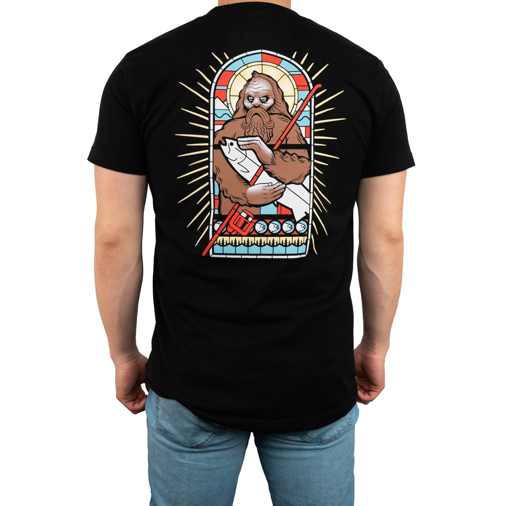 A person wears the STLHD Mens Saint Sasquatch Black T-Shirt by STLHD GEAR, featuring a vibrant stained-glass Bigfoot holding a fish on the back, surrounded by rays and shapes—all crafted in breathable fabric for all-day comfort.