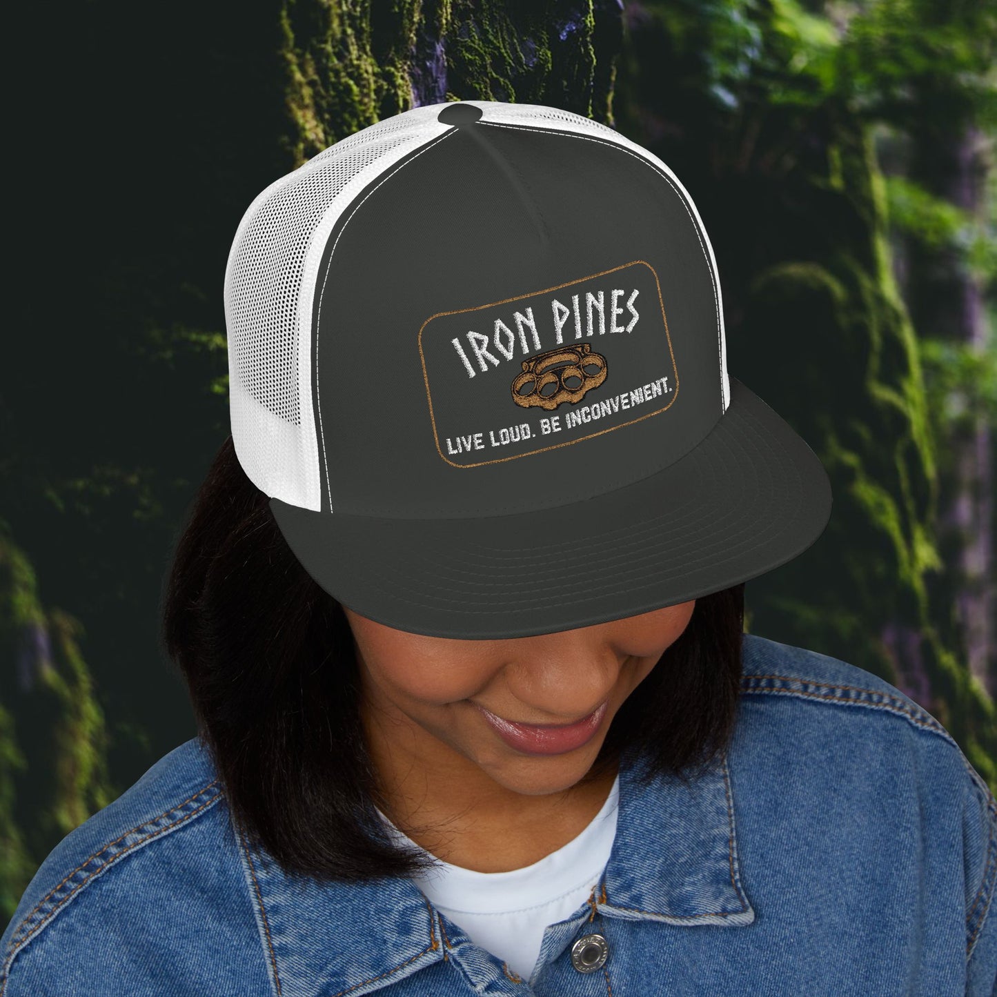 A person smiles while wearing the Printify Iron Pines Trucker Hat – “Live Loud. Be Inconvenient.” Brass Knuckle Edition, featuring a pinecone patch, as the high-profile snapback sits low on their head.