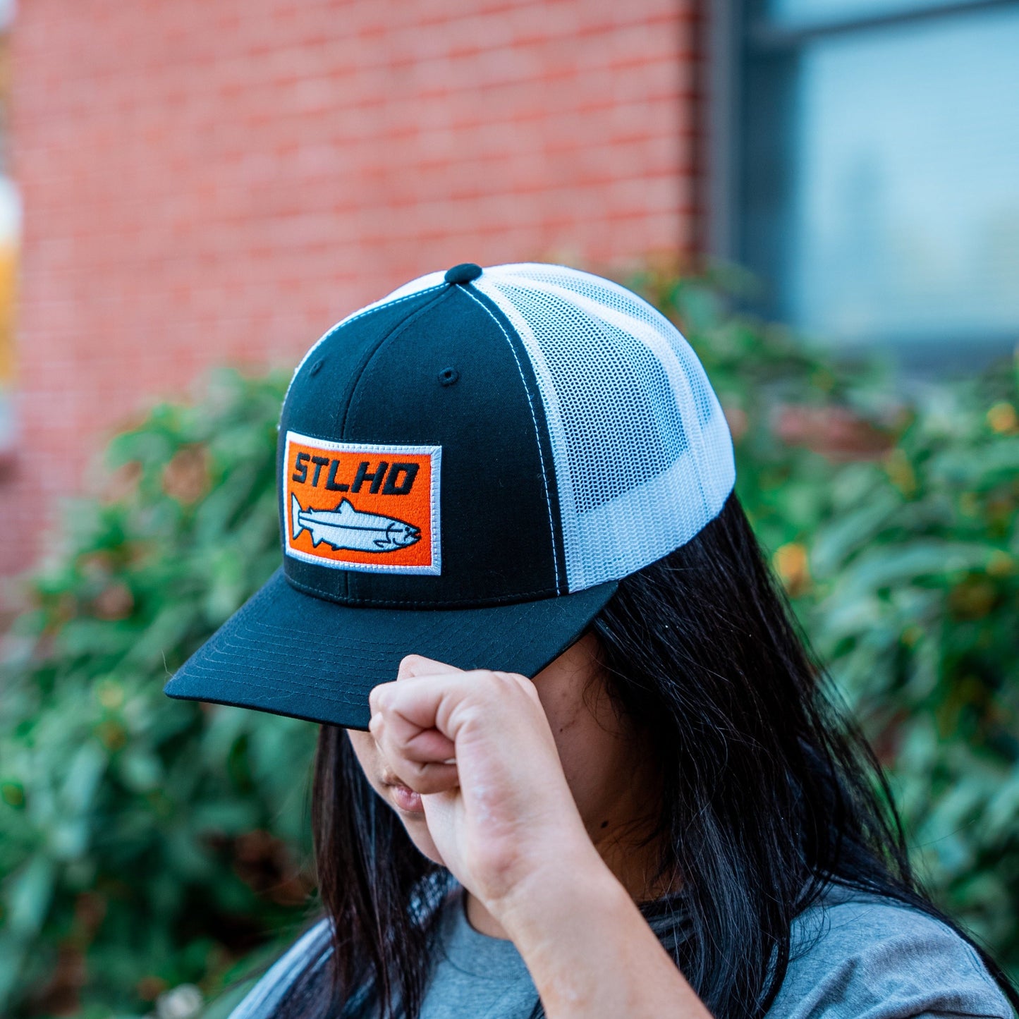 A person with long dark hair tilts the adjustable snapback of an STLHD GEAR Standard White & Black Trucker Hat, featuring an orange fish patch. Their face is partially obscured, with greenery and a brick wall blurred in the background.