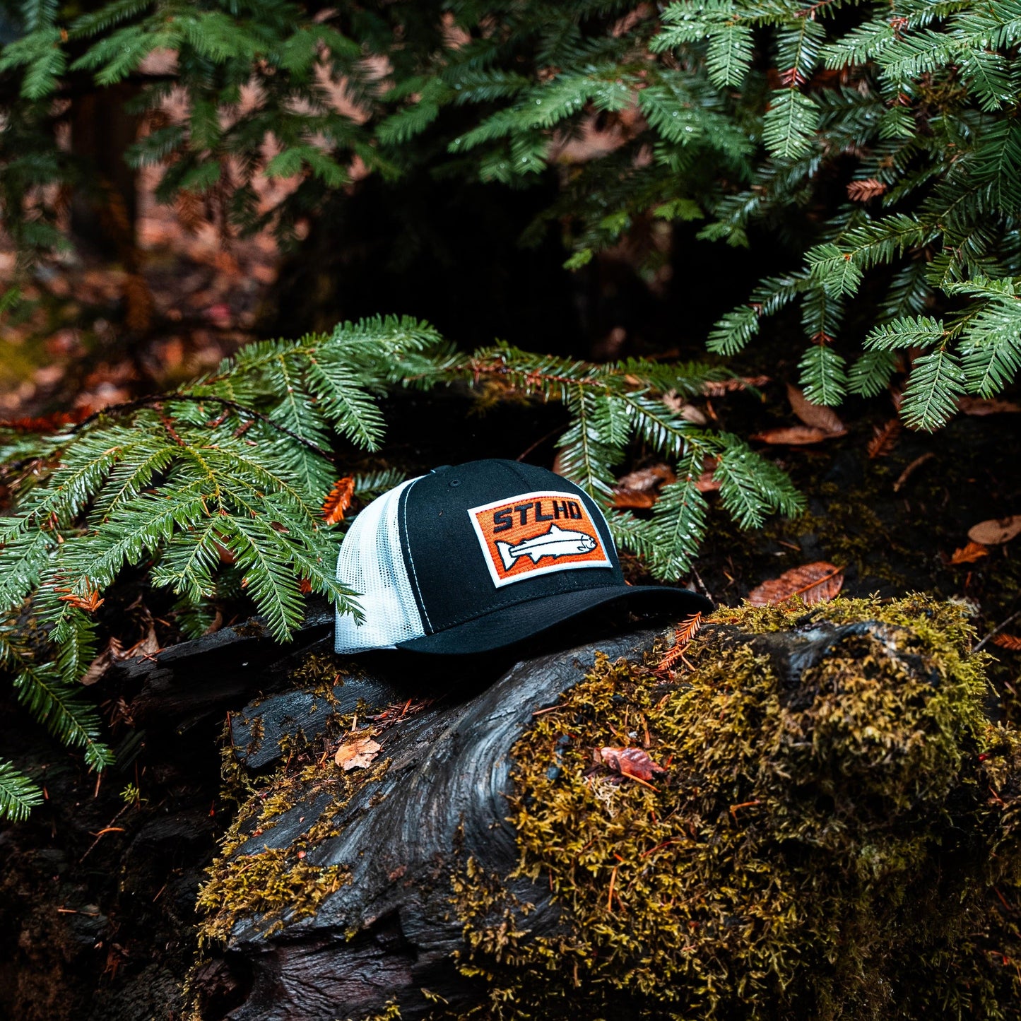 The STLHD GEAR Standard White & Black Trucker Hat, featuring an orange patch with fish graphic and adjustable snapback, rests on a mossy log in a pine forest.
