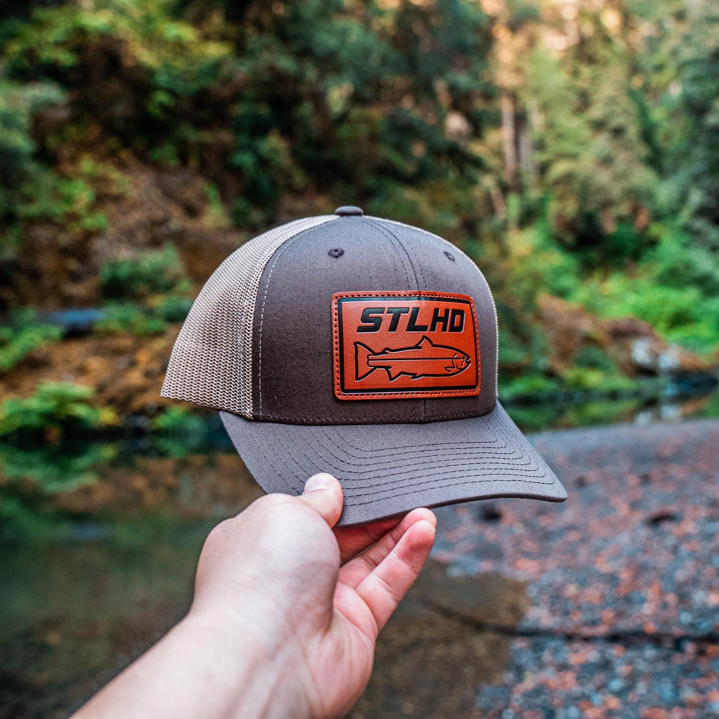 A hand holds the STLHD GEAR Steelhide Brown/Khaki Hat, featuring an orange fish patch, against a blurred backdrop of trees and a rocky creek. This adjustable snapback trucker hat blends rugged style with outdoor spirit.