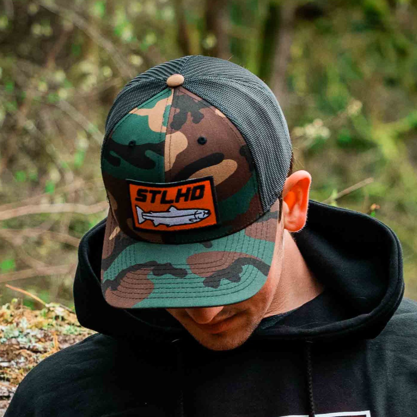Wearing an STLHD GEAR Camo Snapback Trucker Hat and a black hoodie, a person stands outdoors amidst greenery. The hat features a patch with STLHD above a fish illustration.