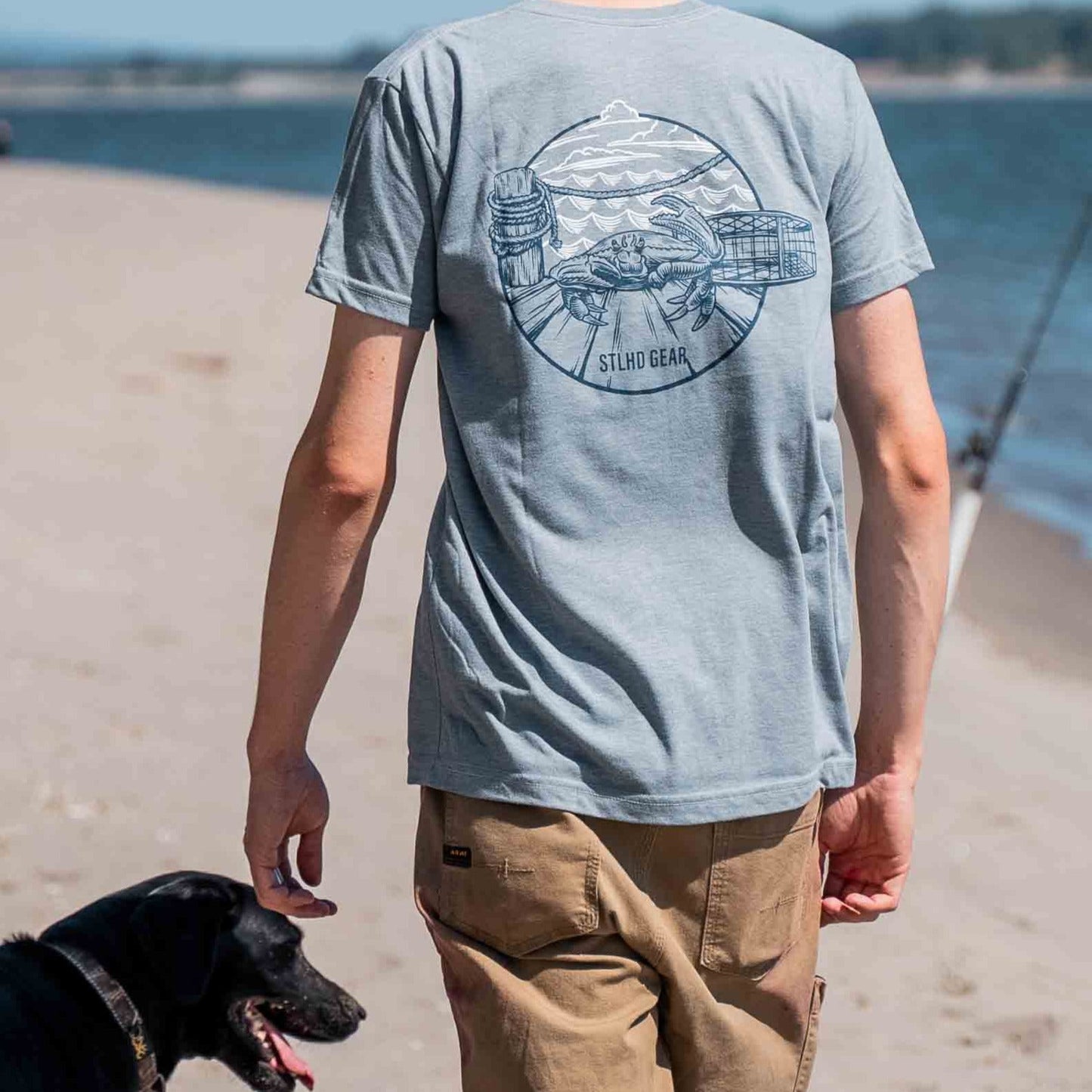 Wearing the STLHD Mens Crabby T-Shirt by STLHD GEAR, a person walks along a sandy beach with a black dog. The light blue shirt features a crab graphic on the back, while the ocean and fishing rod highlight its breathable summer style.