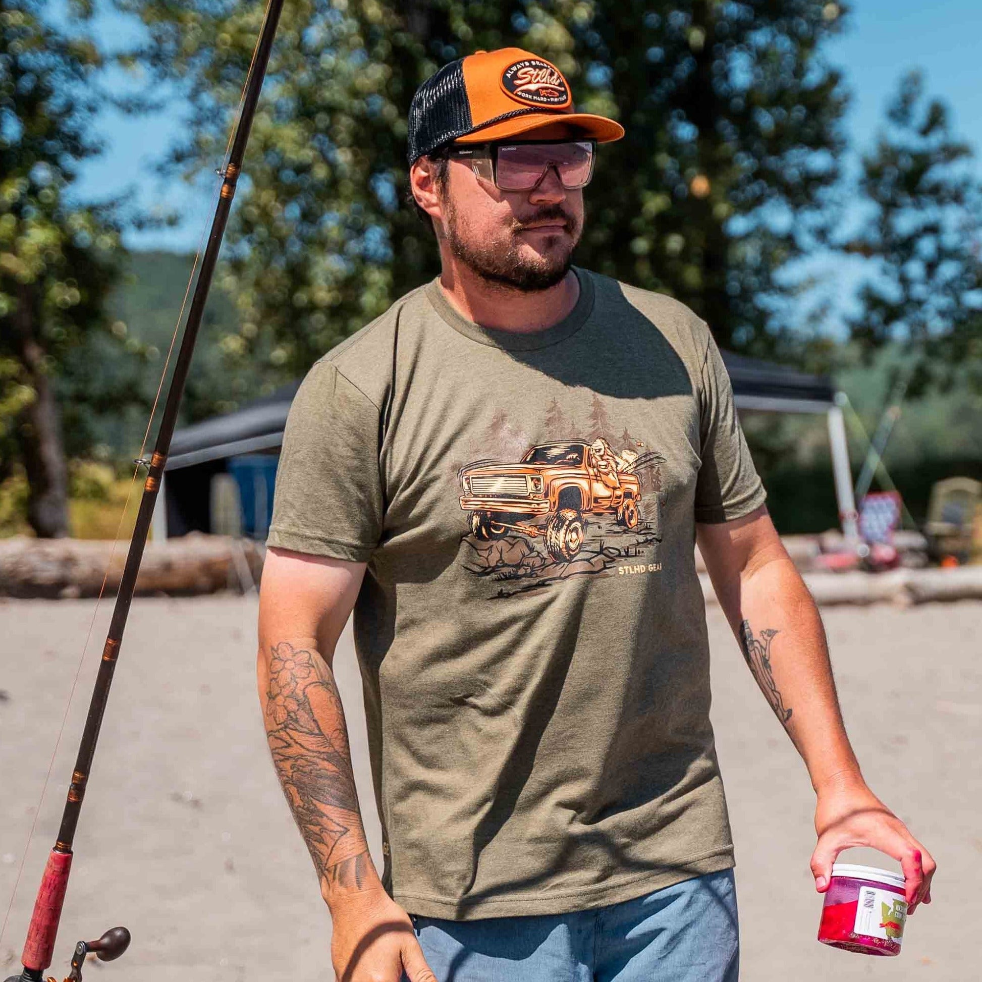 A man in glasses, a trucker hat, and the STLHD GEAR Men’s 4x4 T-Shirt stands outdoors with fishing gear and bait, trees and a tent behind him—an ideal scene for outdoor enthusiasts.
