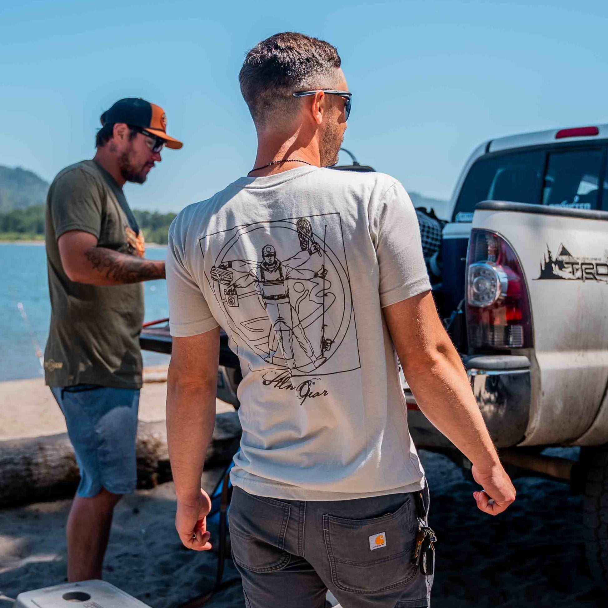 Two men relax lakeside by a white pickup under the sun. One, in sunglasses, wears the STLHD Men’s Anatomy T-Shirt by STLHD GEAR; the other sports a dark shirt and cap. The vibe is casual and outdoorsy.