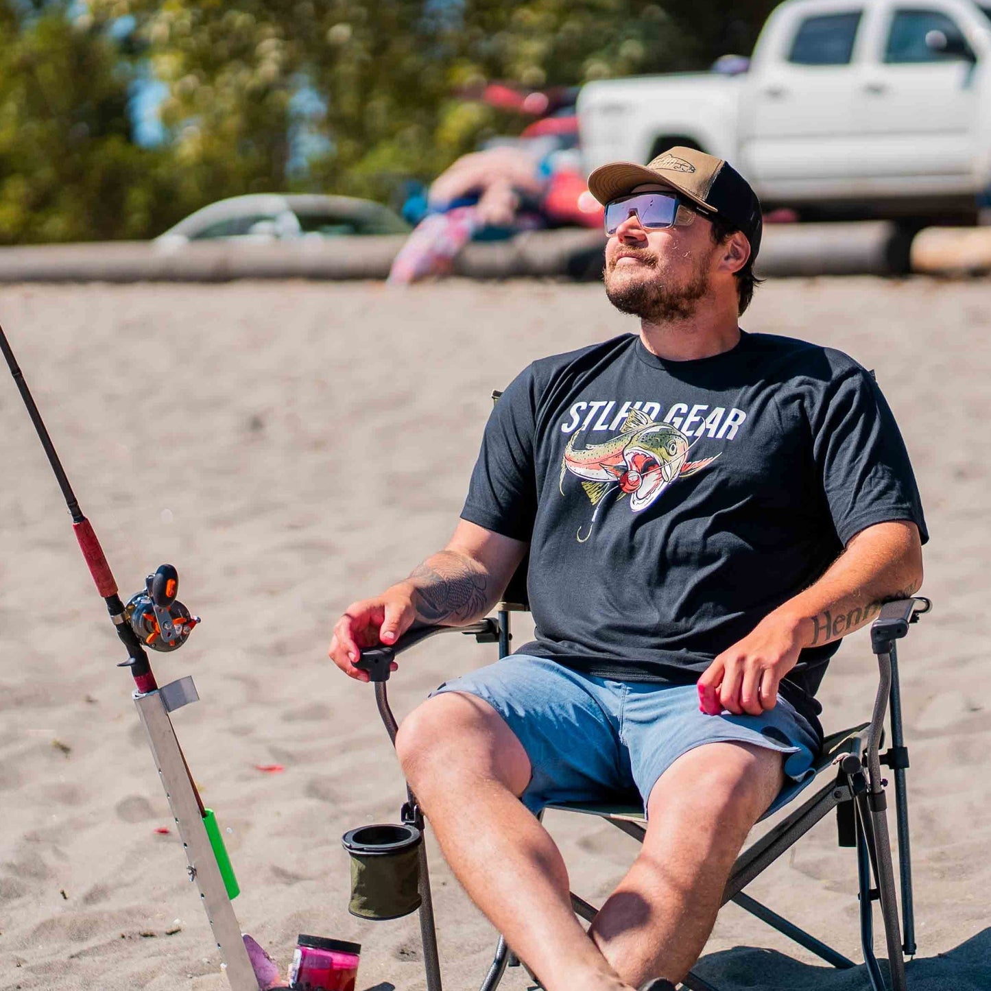 A man in sunglasses and a cap, wearing the STLHD GEAR Men’s Chomper T-Shirt, relaxes on a folding chair at the sandy beach holding a fishing rod. A truck, blurred people, and trees are visible in the background.