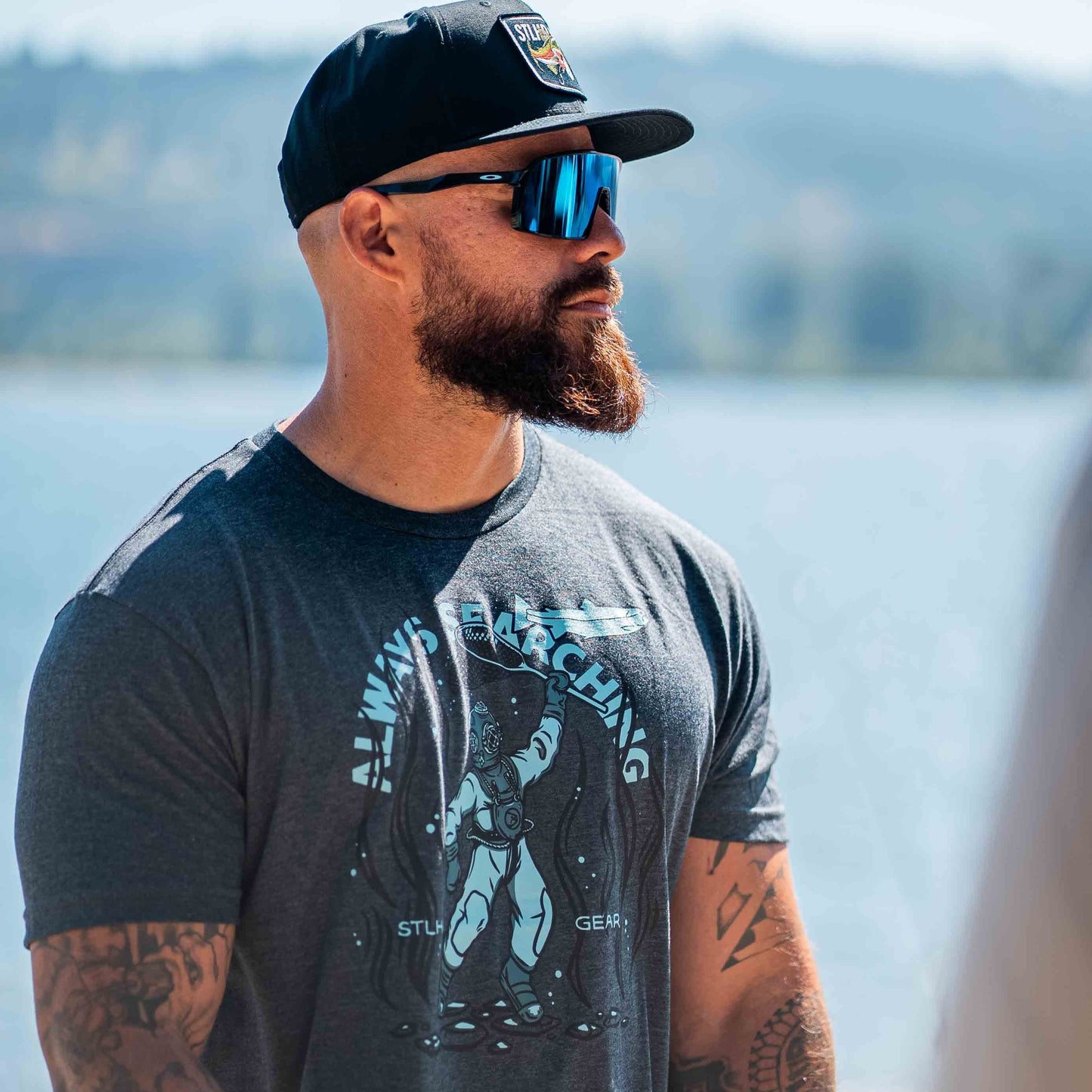 A man wearing sunglasses, a black cap, and the STLHD GEAR Men’s Deep Dive T-Shirt stands by the water with blurred trees and mountains in the background—perfect for any outdoor adventure.