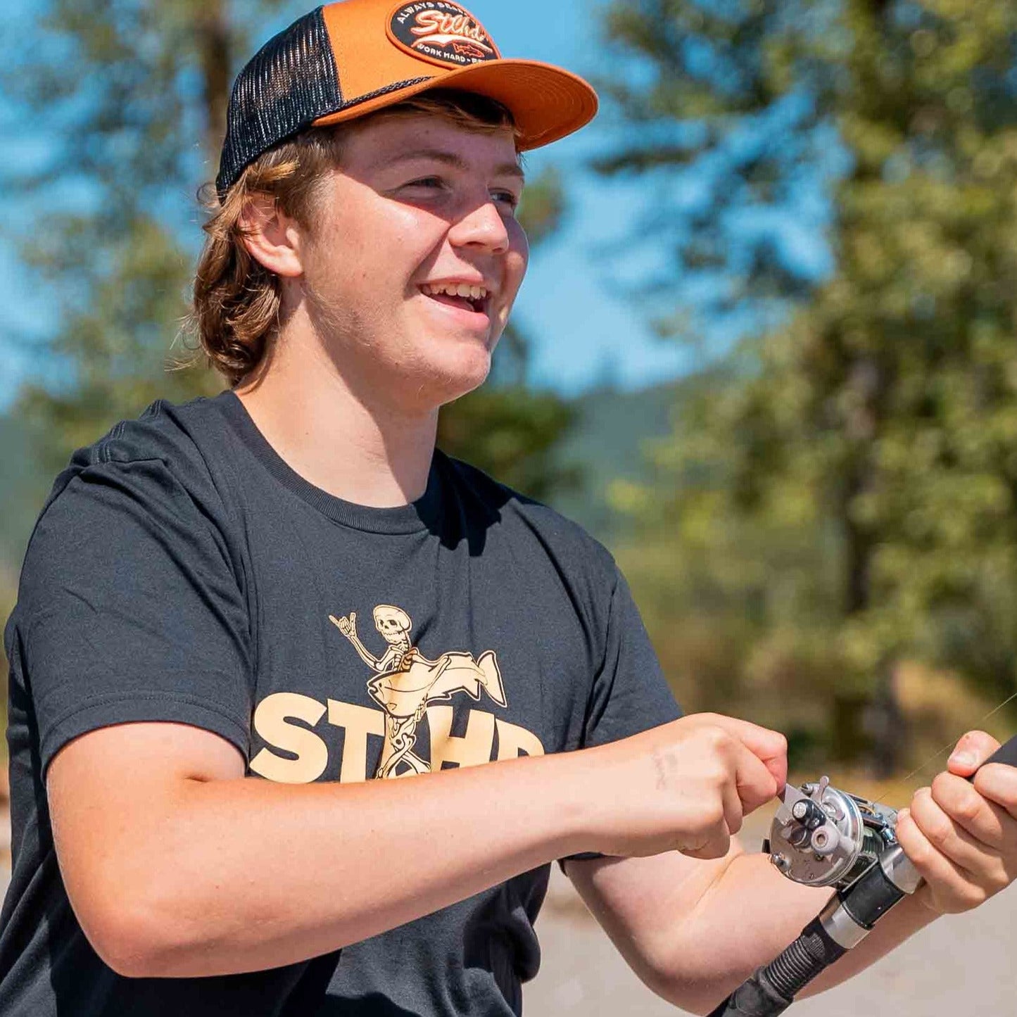 A smiling young person in an STLHD Loosey Goosey T-Shirt by STLHD GEAR and a black-and-orange cap is fishing outdoors on a sunny day, surrounded by trees and a blue sky.