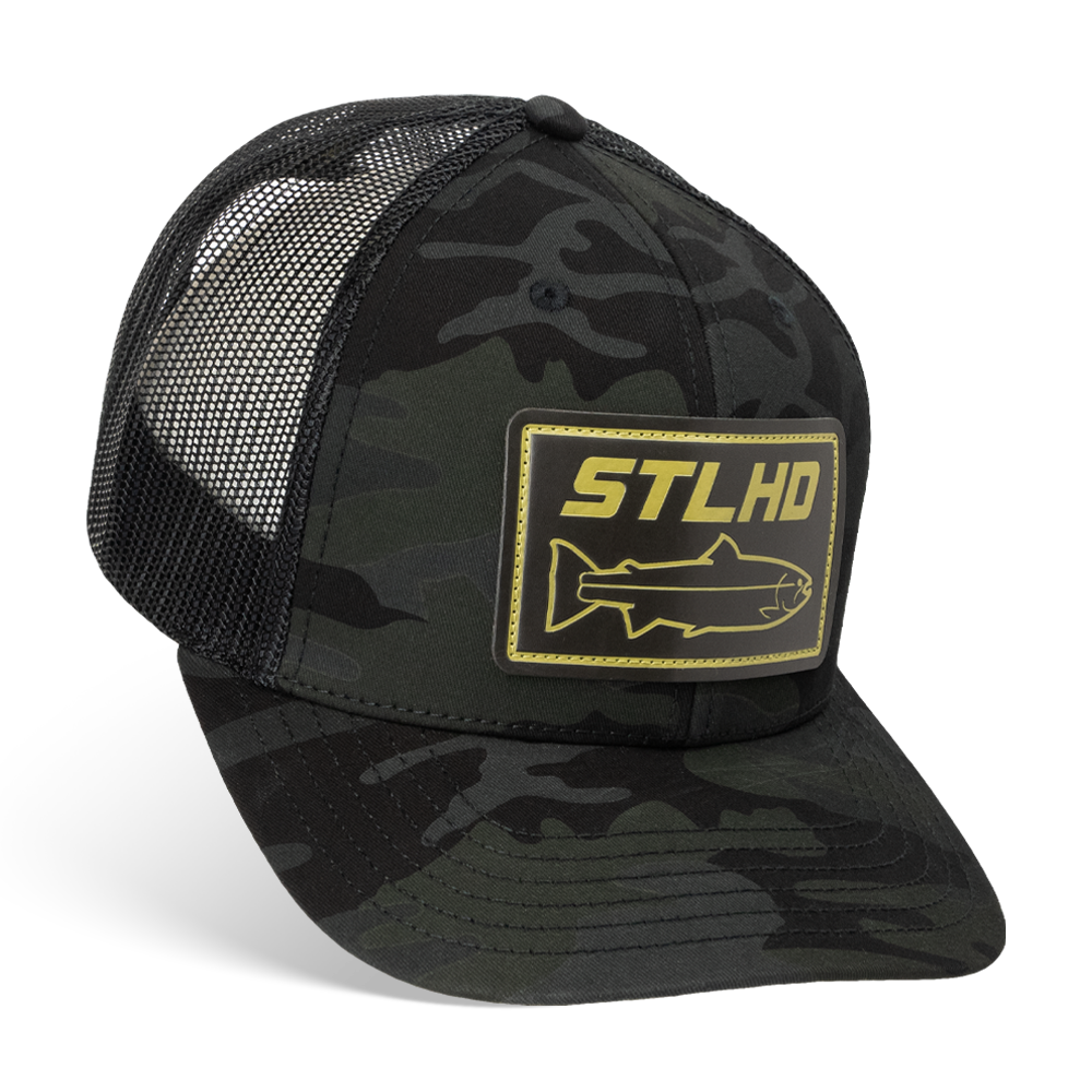 The STLHD GEAR Black Ops Multicam Snapback Trucker Hat features a black and dark camo design, a yellow STLHD fish patch on the front, breathable mesh back, and adjustable snapback for a comfortable fit.