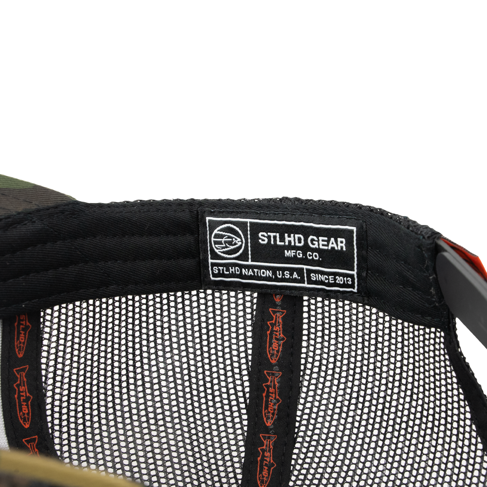 Close-up of the STLHD Camo Snapback Trucker Hat by STLHD GEAR, showing a black label with STLHD GEAR MFG. CO. STLHD NATION, U.S.A. SINCE 2013, orange fish graphics on a mesh interior, and an adjustable snapback for the perfect fit.