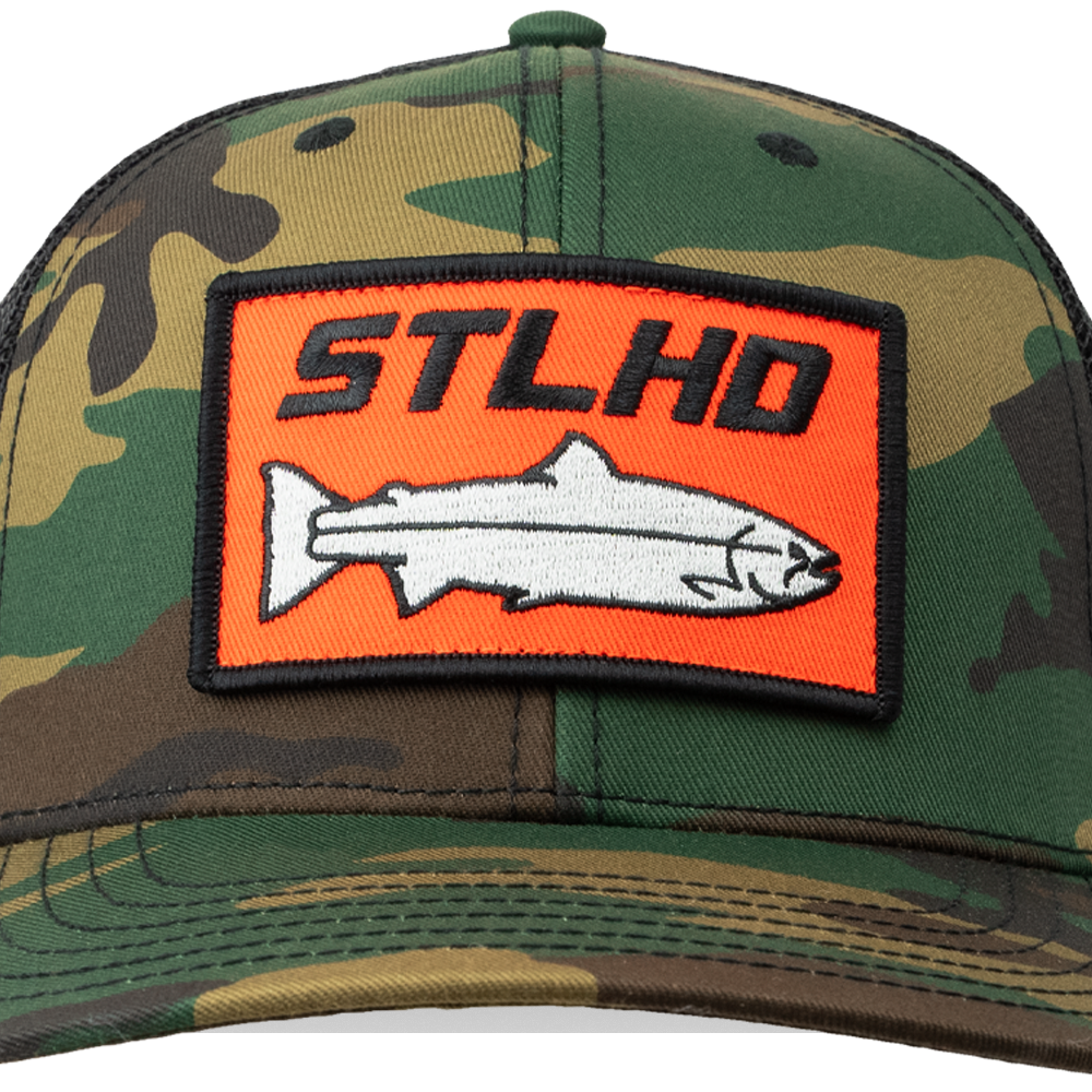 The STLHD GEAR Camo Snapback Trucker Hat features a rectangular orange patch with STLHD above a white fish outlined in black, combining style and function for fishing and outdoor adventures.