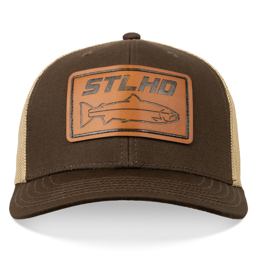 The STLHD GEAR Steelhide Brown/Khaki Hat is a brown and tan leather trucker hat with an adjustable snapback and a front leather patch displaying “STLHD” above a fish outline.