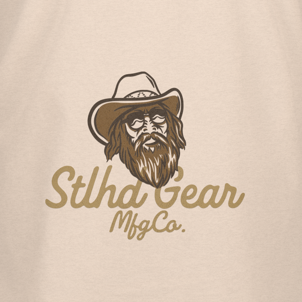 This beige STLHD Country State of Mind T-Shirt by STLHD GEAR features a bearded man in sunglasses and a cowboy hat above “Stlhd Gear Mfg Co.”—a great pick for outdoor enthusiasts and fans of unique graphic tees.